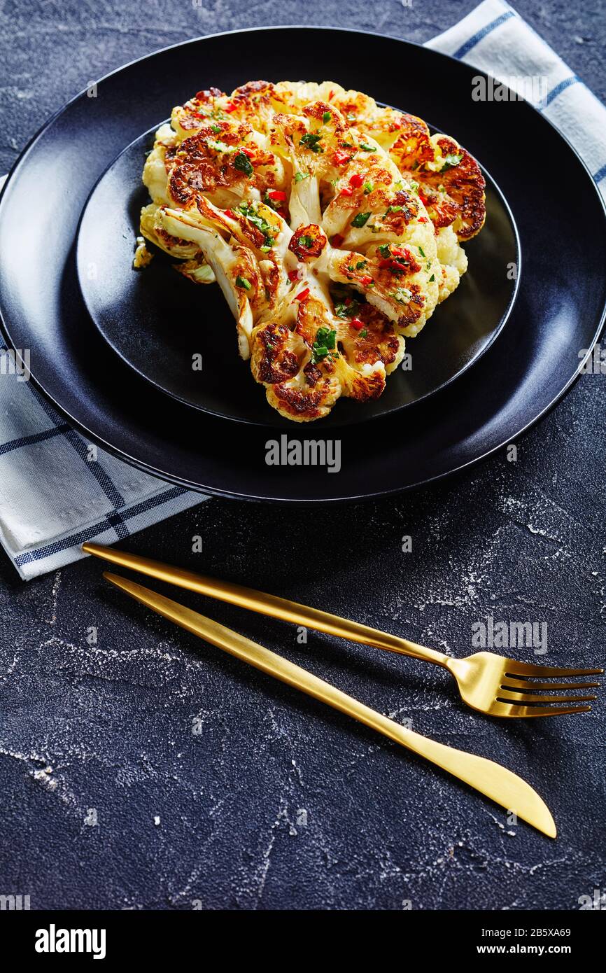 Deliziose bistecche di cavolfiore al forno su un piatto nero cosparso di salsa di peperoncino rosso, aceto, aglio e coriandolo, servito con oro Foto Stock