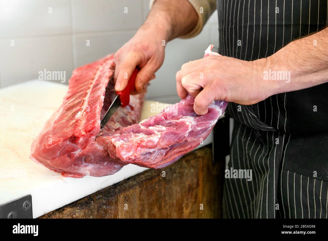 Macellaio affettare un filetto di maiale crudo fuori dall'osso con un coltello affilato su un blocco macellaio in un primo piano sulle sue mani Foto Stock