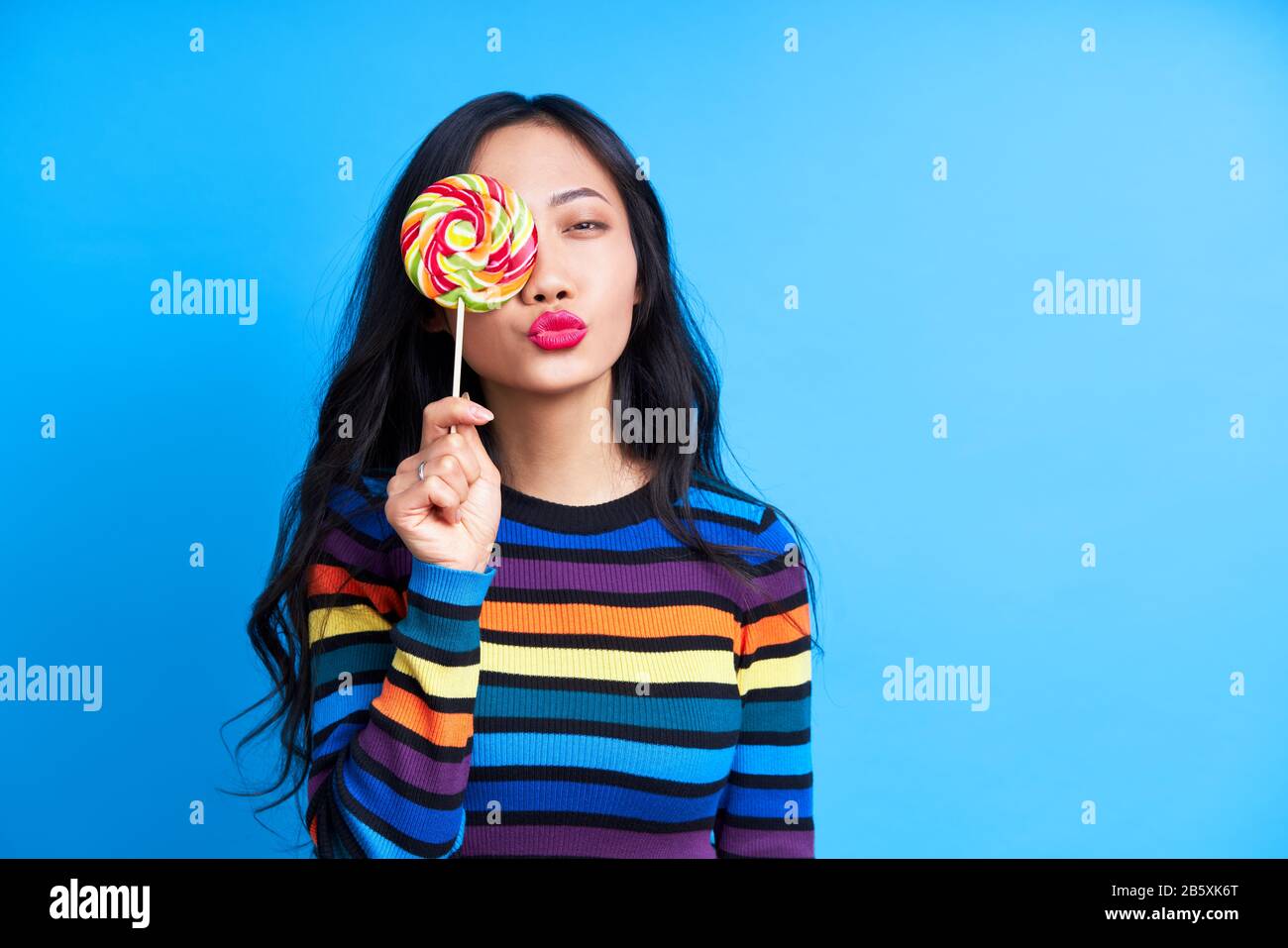 Bella donna inviare aria bacio e coprire il suo occhio con lollipop colorato isolato su sfondo blu. Concetto di emozioni, cibo dolce Foto Stock