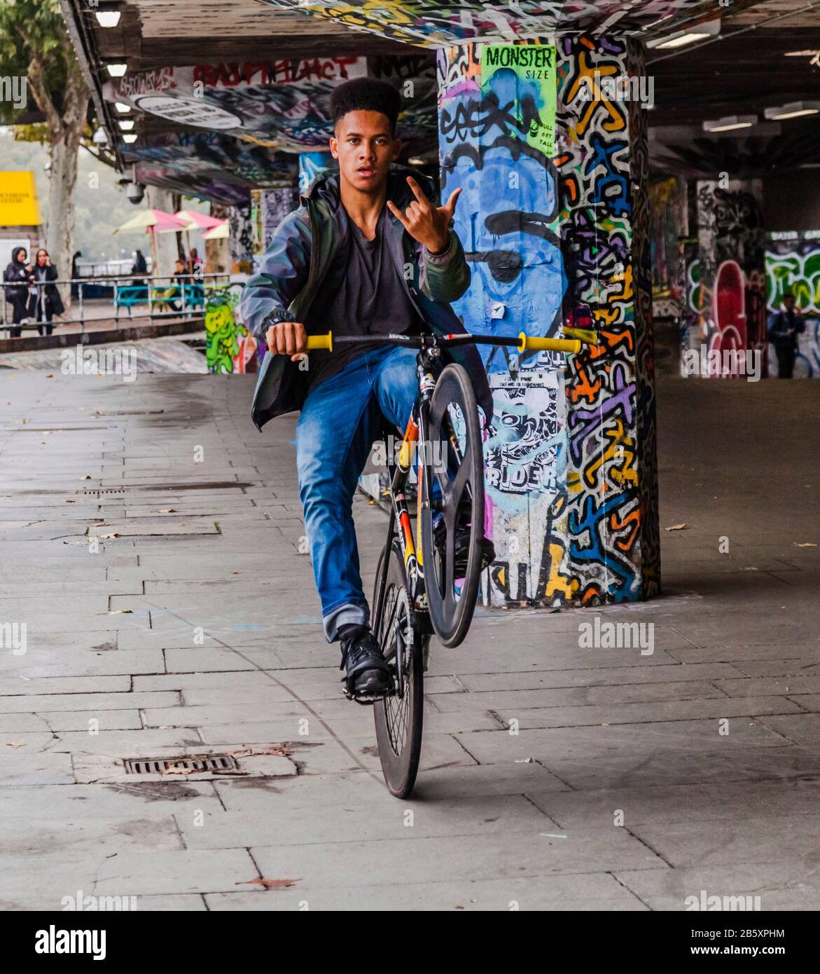 Un giovane nero che guida la sua moto su una ruota fa gesto con le dita verso la macchina fotografica all'Undercroft Skate Park in South Bank, Londra, Inghilterra, Regno Unito Foto Stock