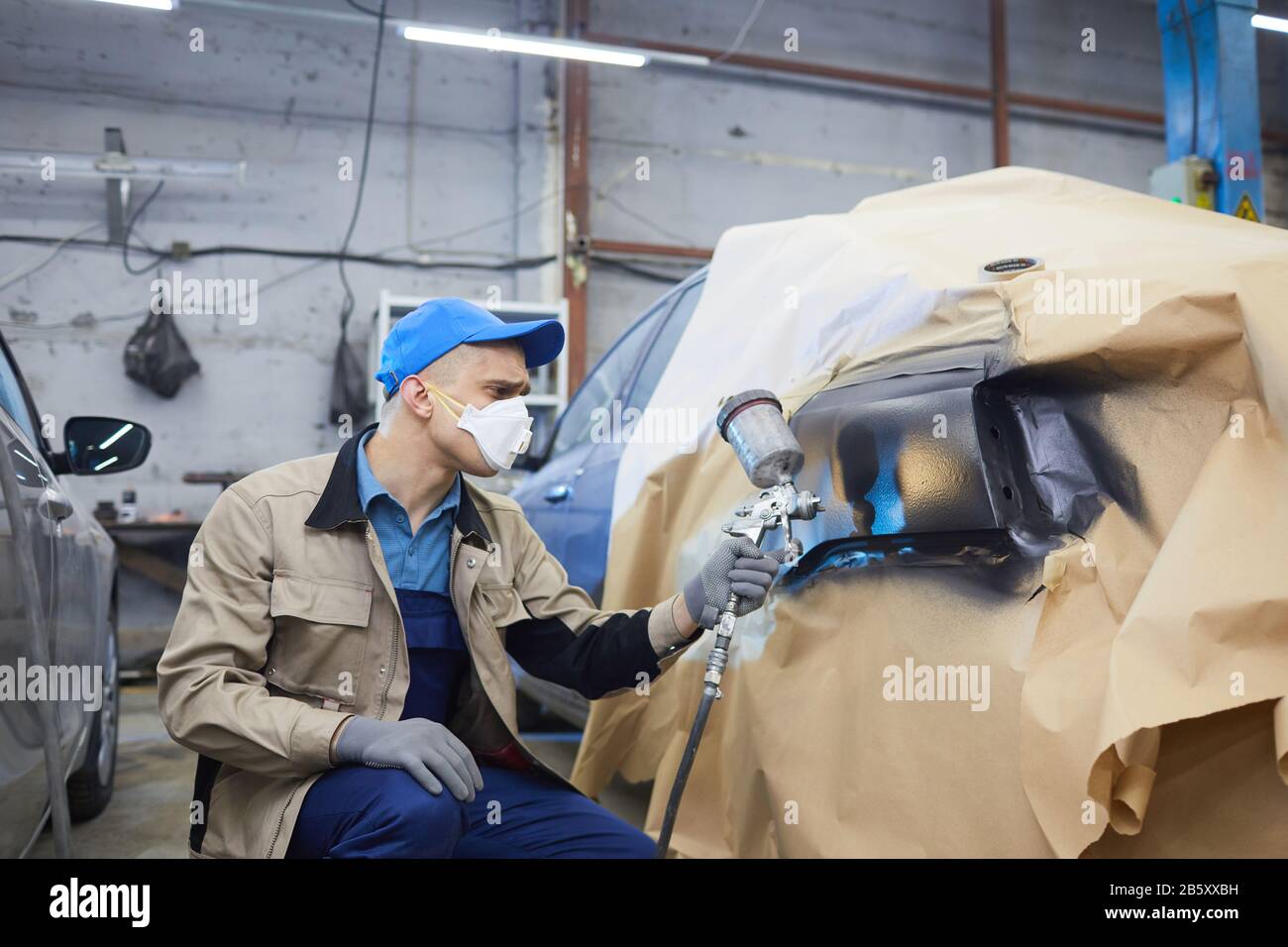 Lavoratore di servizio auto non riconoscibile che indossa uniforme e maschera respiratore sulla vettura di verniciatura a viso con pistola a spruzzo, colpo orizzontale Foto Stock
