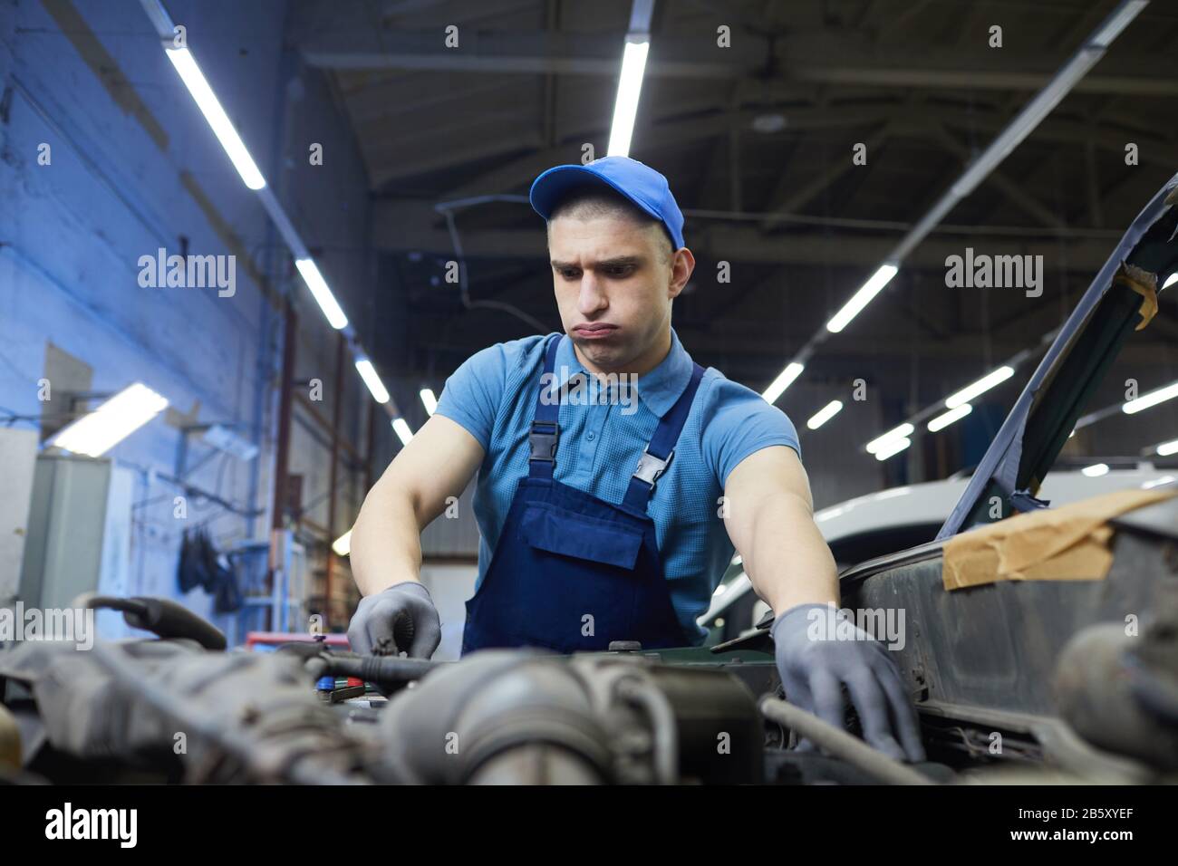 Ritratto di meccanico automatico concentrato con guance gonfiate riparazione motore auto, orizzontale medio ritratto Foto Stock