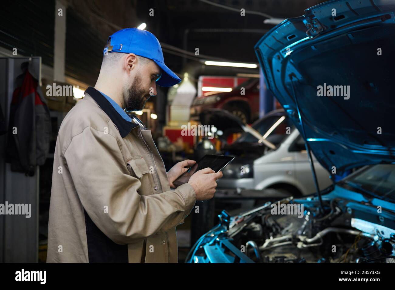 Uomo Cuacsian con barba sul viso che lavora in officina per l'assistenza automatica utilizzando un tablet PC, scatto orizzontale con vista laterale Foto Stock