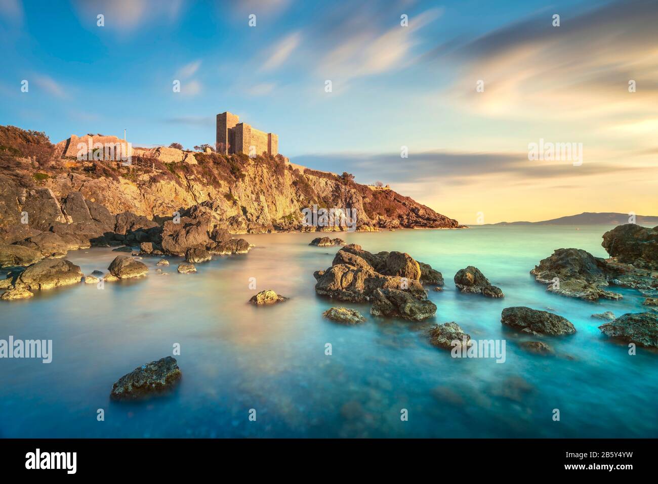 Spiaggia rocciosa di Talamone e fortezza medievale Rocca Aldobrandesca e mura al tramonto. Maremma Argentario destinazione turistica italiana. Toscana, Italia. Foto Stock