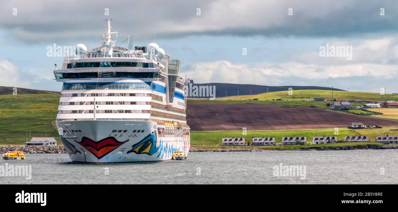 Nave da crociera tedesca AIDAbella, ormeggiata al largo di Lerwick, Shetland torri sopra case vicine sulla riva. Gestito da AIDA Cruises, parte di Carnival Corporation. Foto Stock