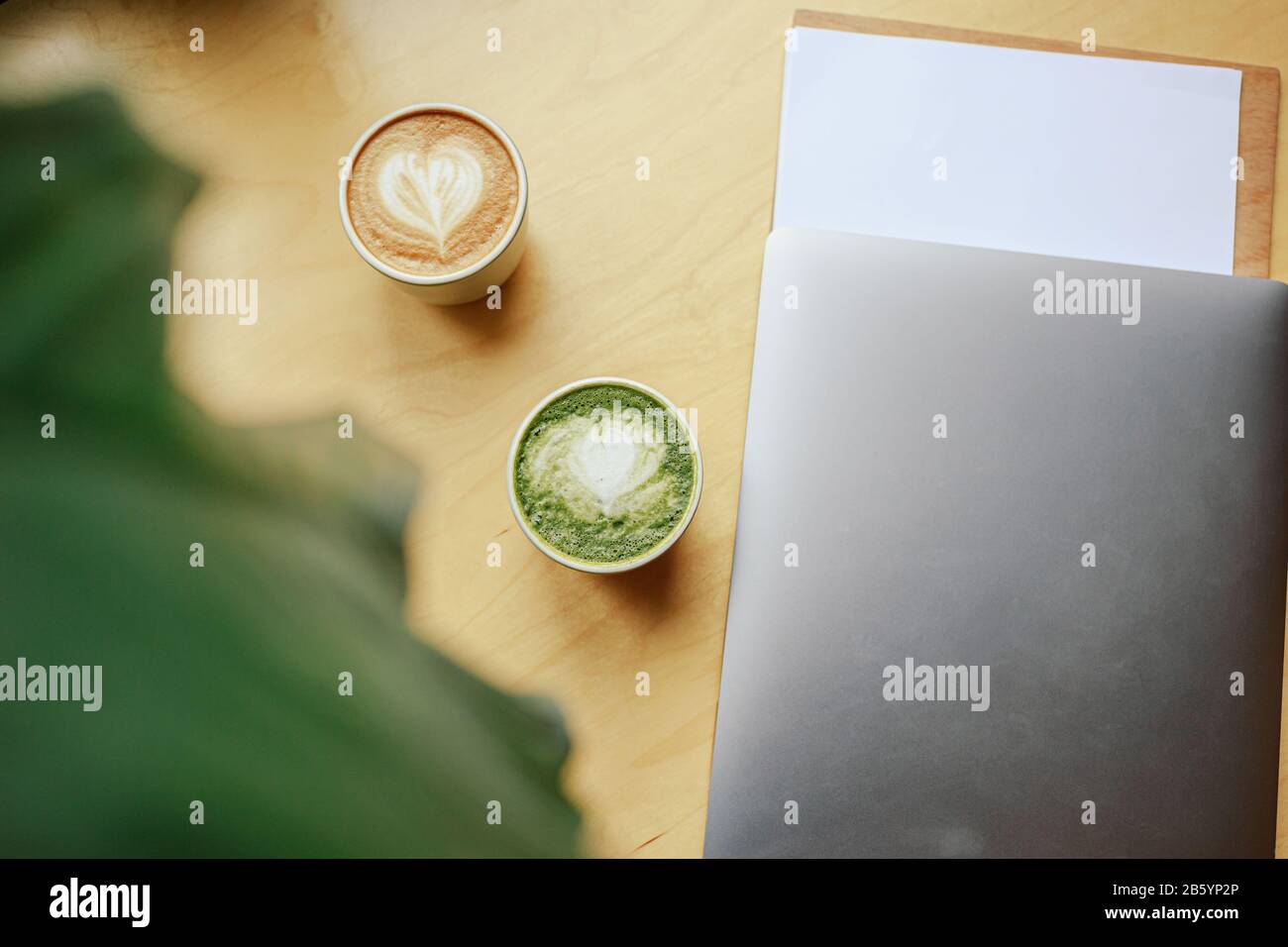 Colazione con due tazze calde con latte art caffè e matcha latte tè e computer portatile in background su tavolo in legno. Disposizione piatta. Spazio di copia. Il conc Foto Stock