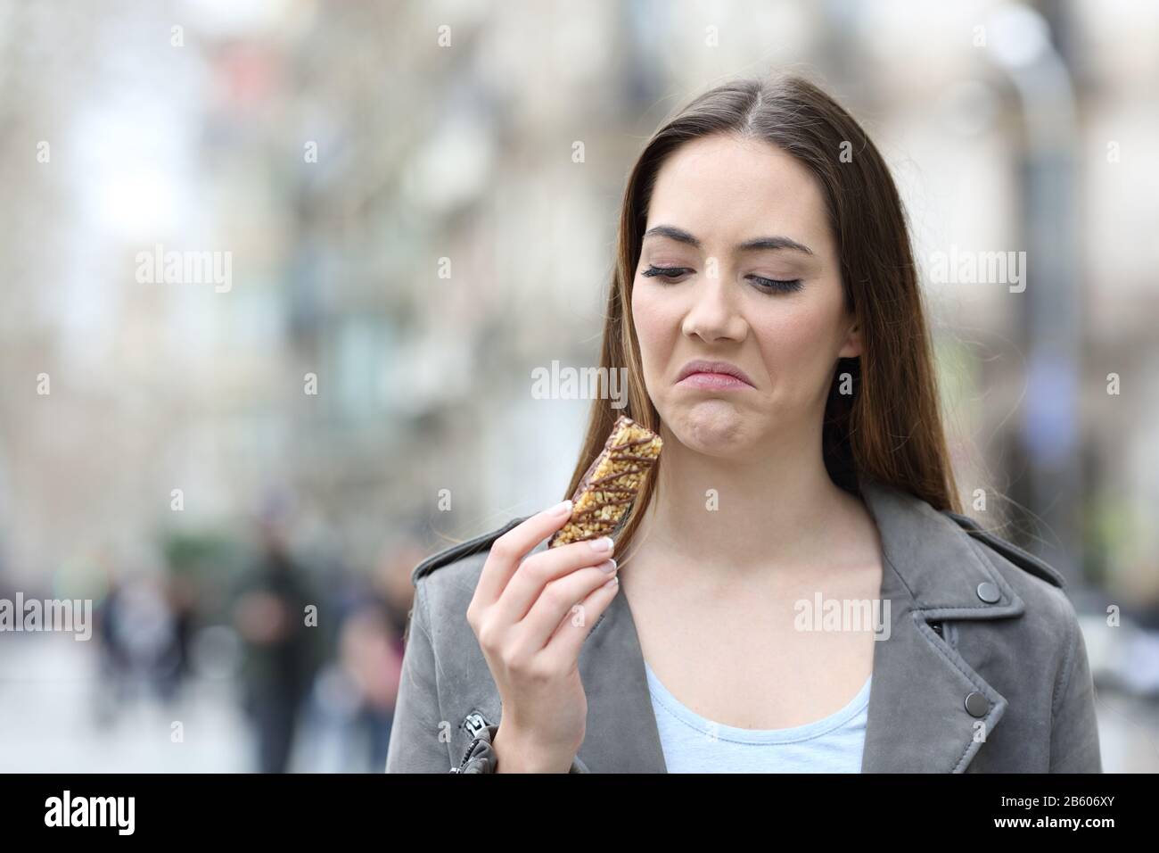 Vista frontale ritratto di una donna disgustata che guarda uno snack cereale bar su una strada della città Foto Stock