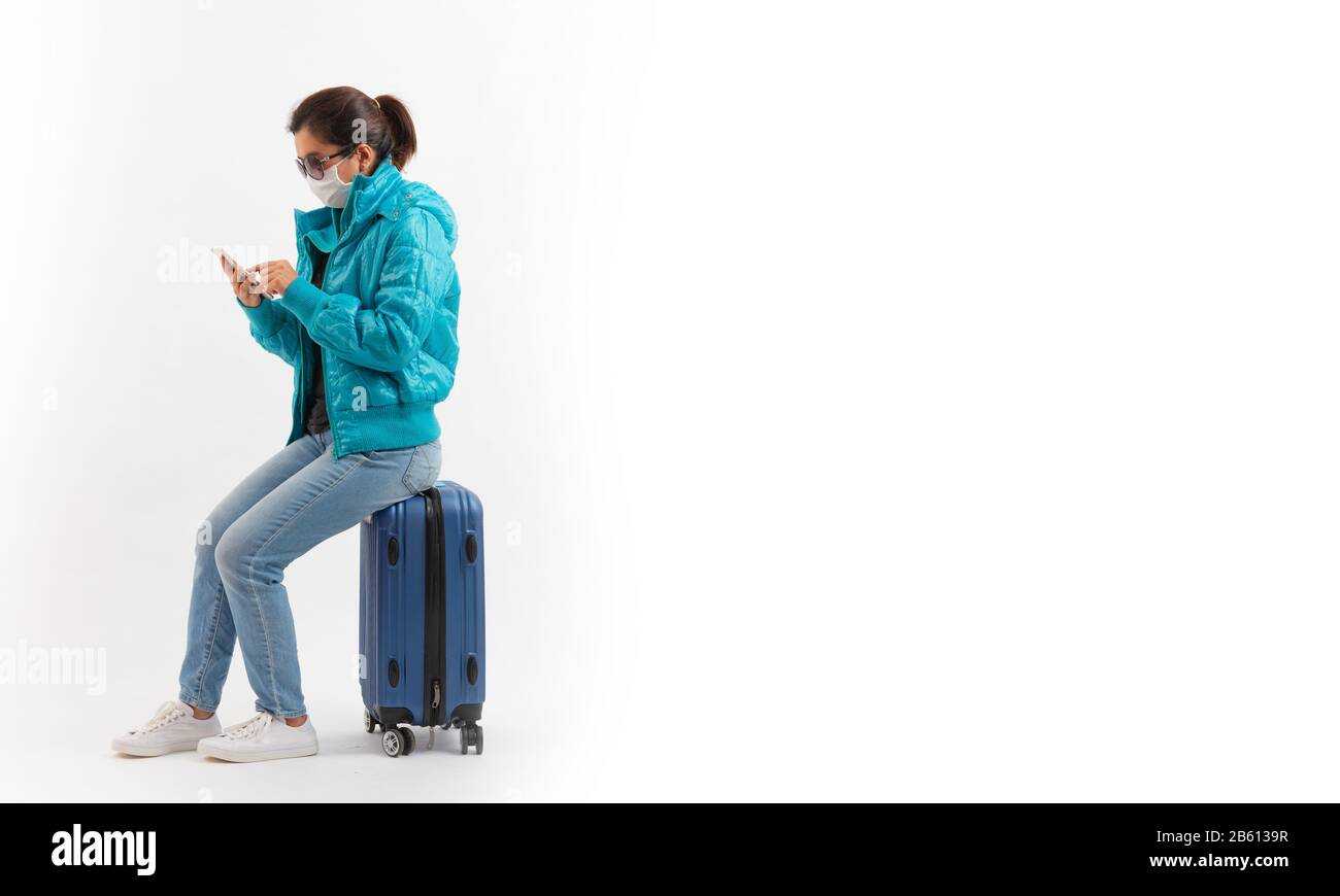 Un turista in maschera medica sedersi sul bagaglio e utilizzando il telefono cellulare isolato su sfondo bianco . Nuovo focolaio di coronavirus COVID-19, Persona presen Foto Stock