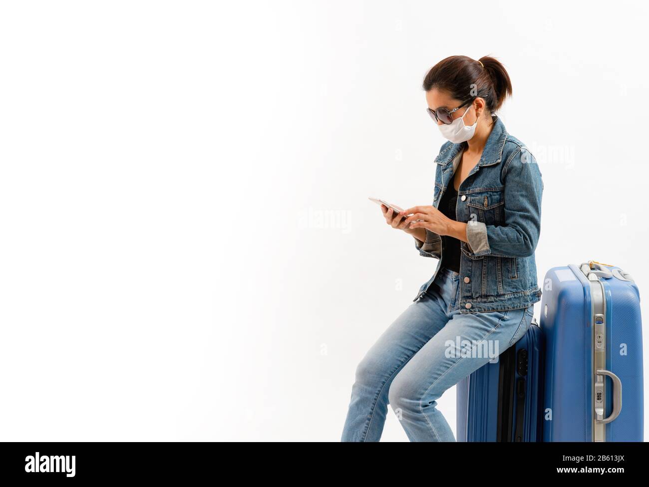Un turista in maschera medica sedersi sul bagaglio e utilizzando il telefono cellulare isolato su sfondo bianco . Nuovo COVID-19 coronavirus focolaio, Persona prevenire Foto Stock