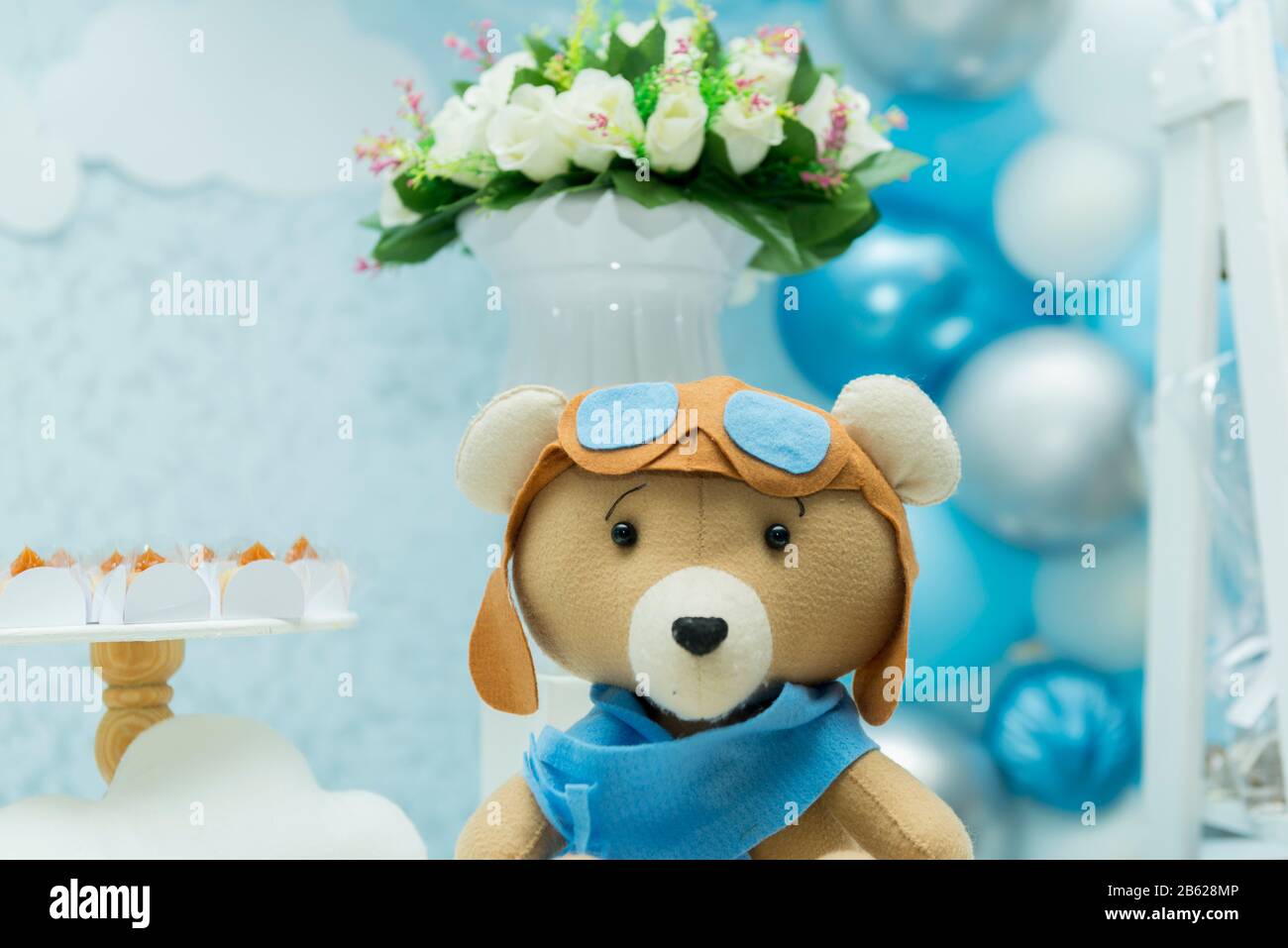 Orsacchiotto con cappello e occhiali da aviatore e un bellissimo bouquet e palloncini blu e bianchi sullo sfondo. Orso con gli occhiali e l'aeroplano d'annata Foto Stock