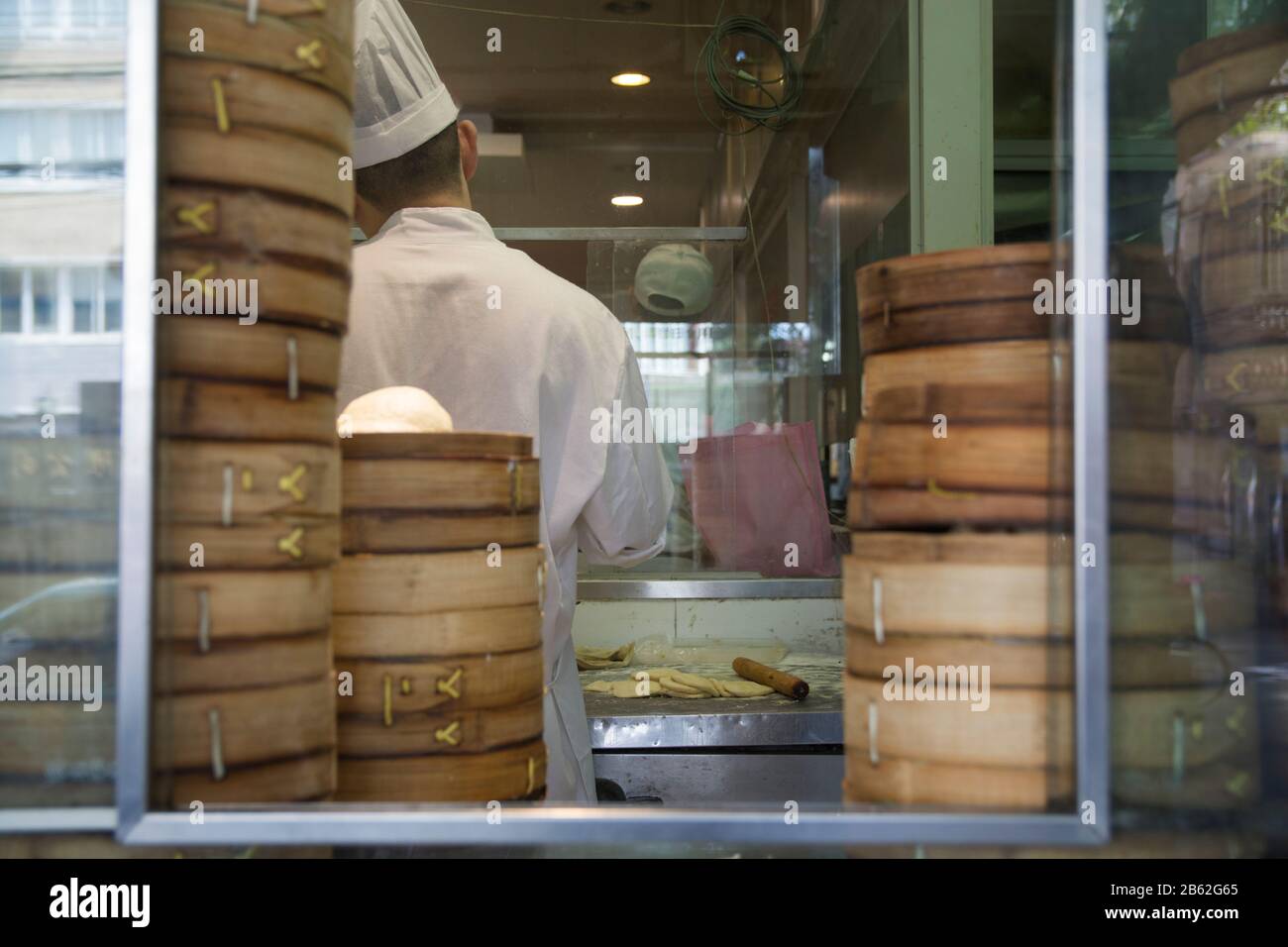 Chef cinese in piedi in una cucina da forno, circondato da gnocchi di scatole di bambù, visto attraverso una finestra. Foto Stock