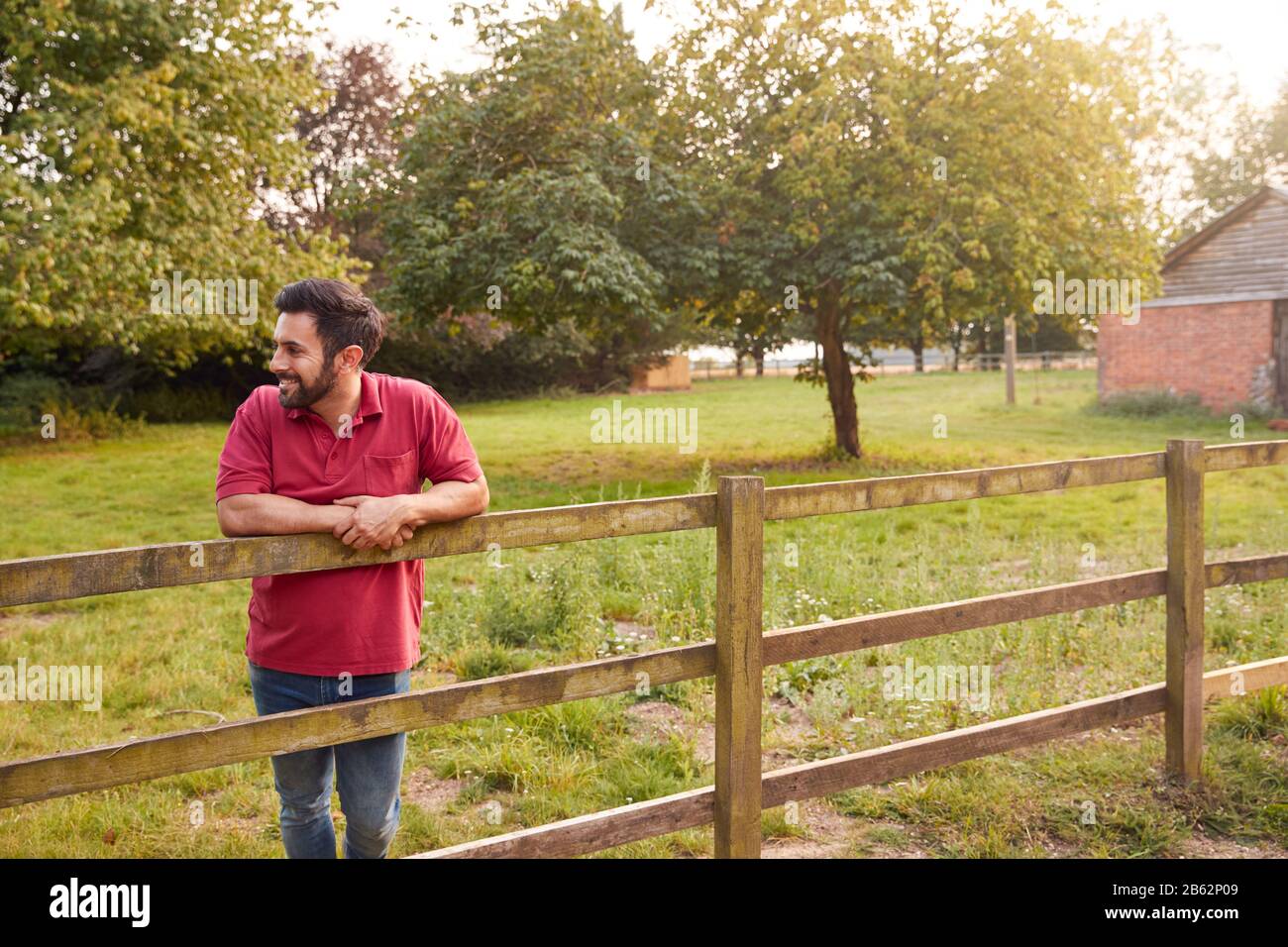 Uomo Sorridente Che Prende Una Pausa E Riposa Su Fence Durante La Passeggiata In Campagna Foto Stock