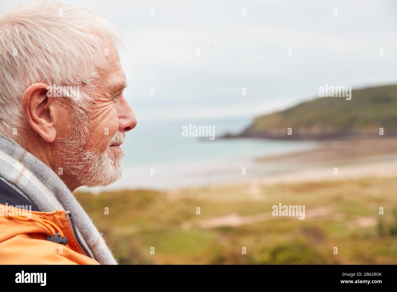 Profilo Shot Of Active Senior Man Passeggiate Lungo Il Percorso Costiero In Inverno Con La Spiaggia E Le Scogliere Dietro Foto Stock