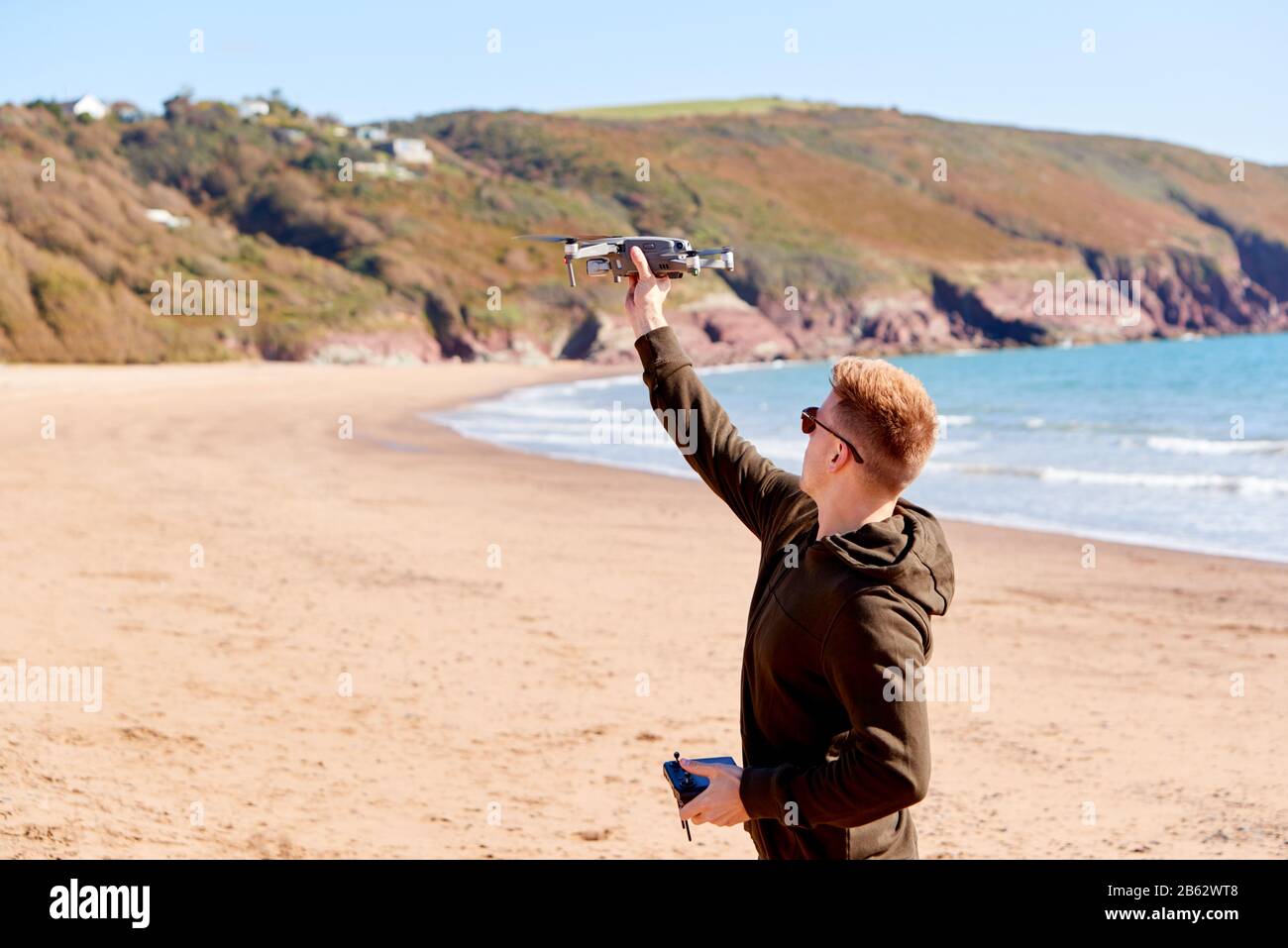 Young Man Flying Drone Con Fotocamera Sulla Spiaggia Con Onde In Background Foto Stock