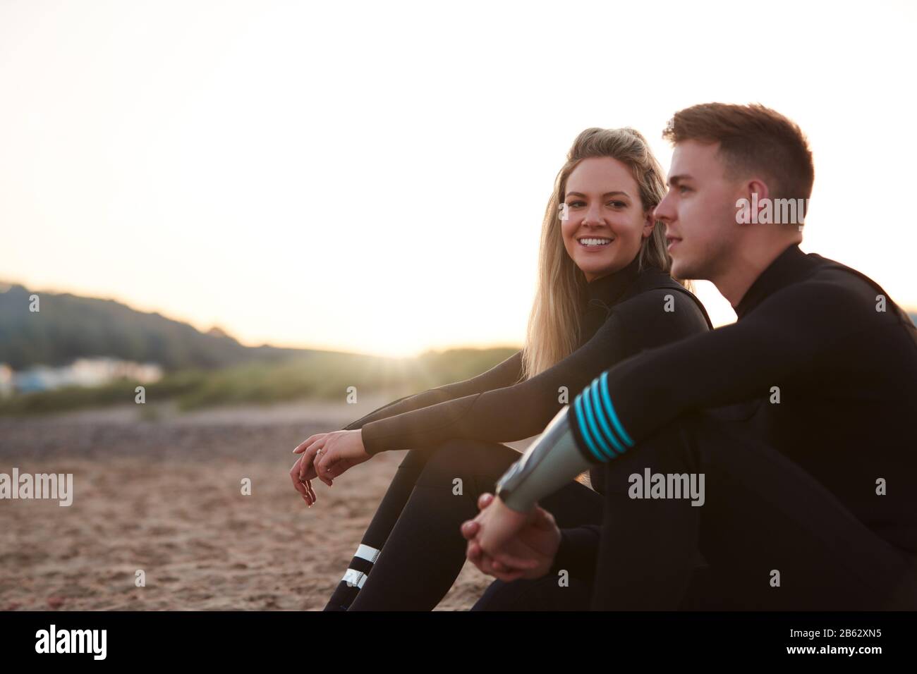 Profilo Vista Di Coppia Indossa Le Mute Su Staycation Surfing Seduto Sulla Spiaggia Che Si Affaccia Sul Mare Foto Stock