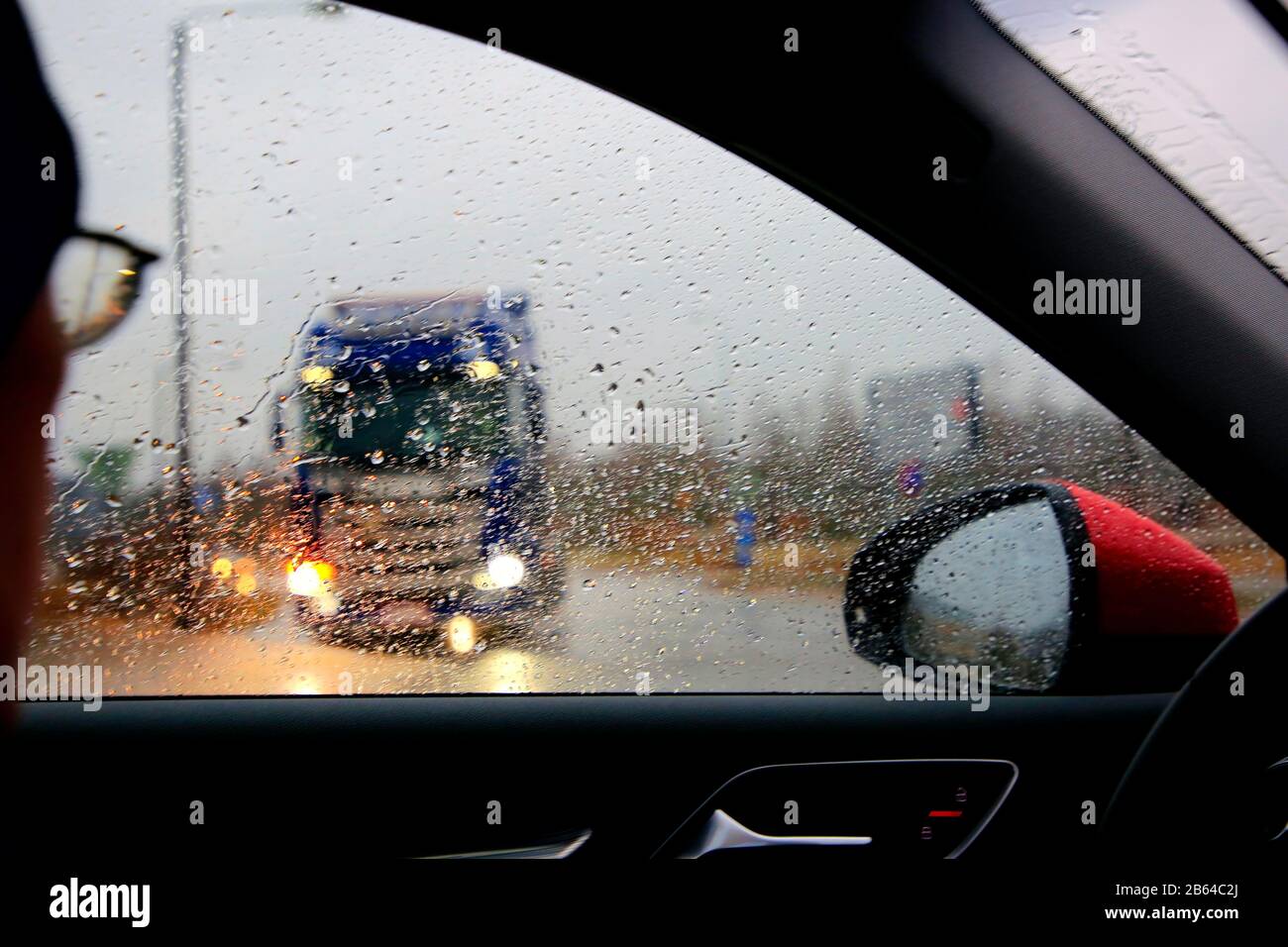 Guida su strada in forte pioggia, conducente che guarda il traffico intersecante con un camion in attesa di entrare in strada. Foto Stock