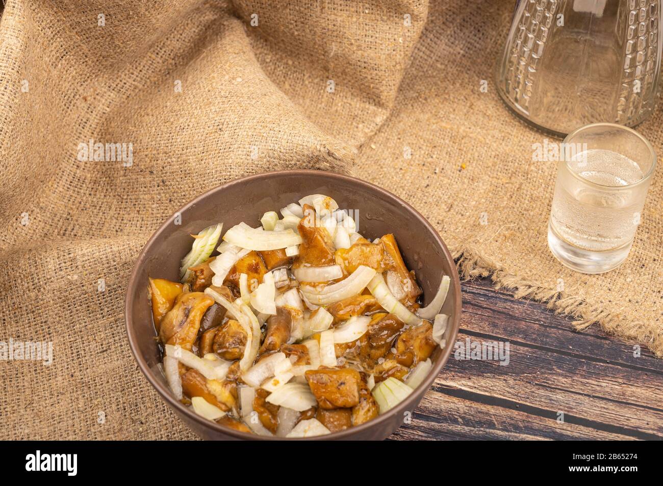 Funghi salati con cipolle in una ciotola di ceramica, un bicchiere di vodka e un decanter su uno sfondo di tessuto di homespun ruvido. Primo piano Foto Stock