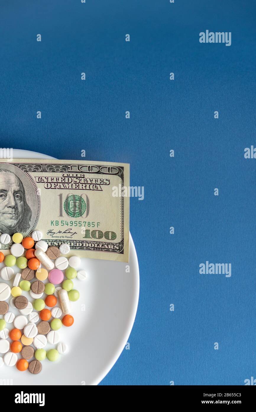 I farmaci costano denaro. Drugs and dolls.White Plate con pillole e capsule multicolore e una fattura da 100 dollari su un classico sfondo blu. Pillole Foto Stock