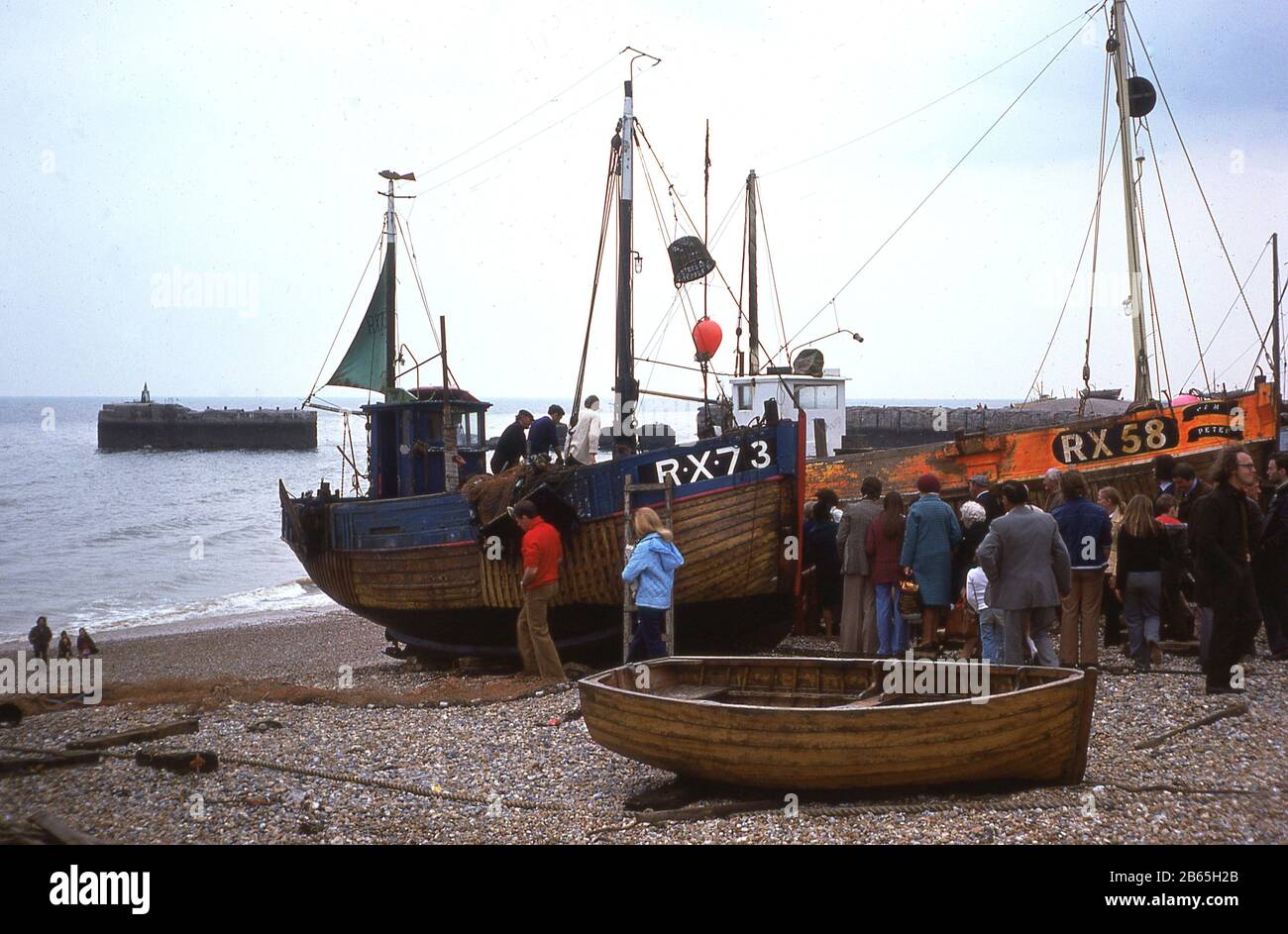 Anni '1970, storia, gente che aspetta a bordo di vecchie barche da pesca in legno sulla spiaggia di stoney a Rock-a-Nore a Hastings, East Sussex, Inghilterra, Regno Unito, la RX73 dipinta di blu e la RX58 dipinta di arancione. Uno dei porti di pesca più antichi della Gran Bretagna, la città di mare ospita la famosa flotta di pesca Hastings, la più antica e grande flotta di pescatori lanciata sulla spiaggia della Gran Bretagna. Foto Stock