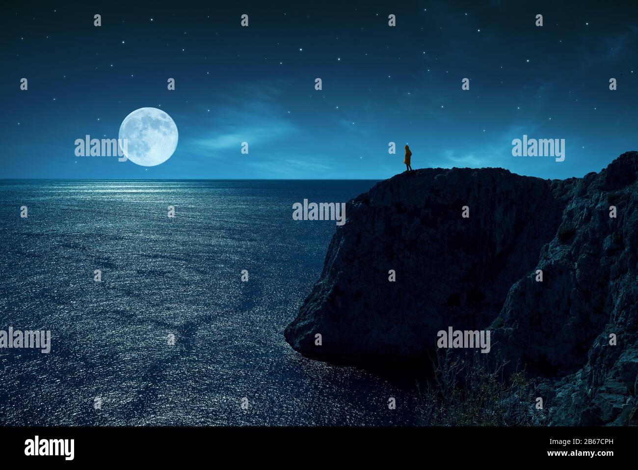 La persona è in piedi nel bordo di una scogliera contro il mare e la luna piena, sotto le stelle e la luce della luna Foto Stock