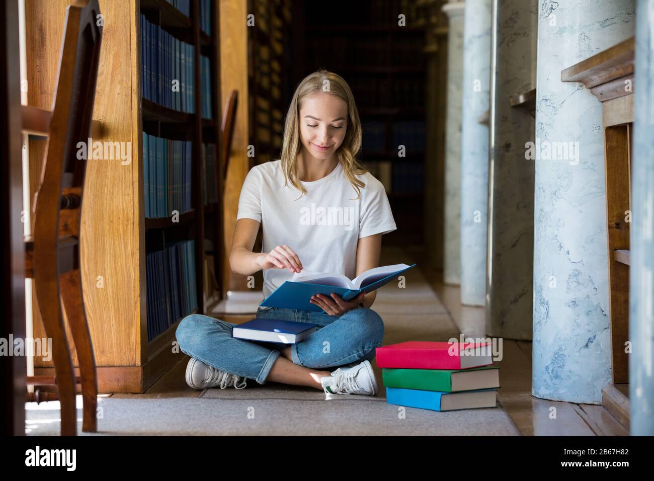 Giovane ragazza seduta in vecchia biblioteca tradizionale a scaffali, alla ricerca di libri.. Sorridente studente che lavora, studia. Istruzione superiore. Foto Stock