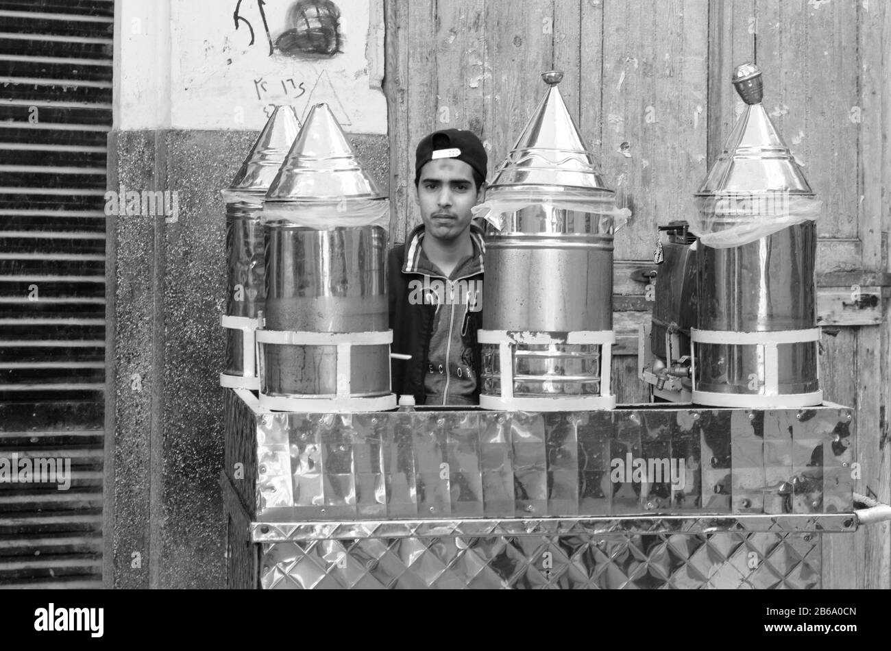 In un'immagine monocromatica che mette in risalto la vita urbana, un giovane venditore si trova tranquillamente dietro il suo carrello in metallo, rivestito di lucido Foto Stock