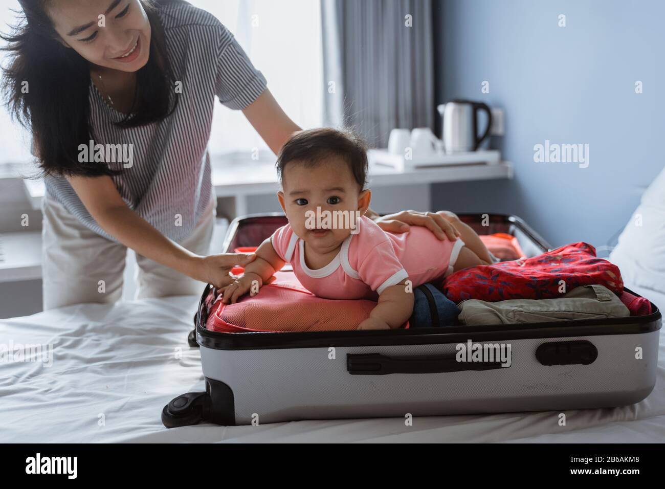 La mamma sorride tenendo un bambino carino sdraiato in una valigia aperta piena di vestiti posti sul letto Foto Stock