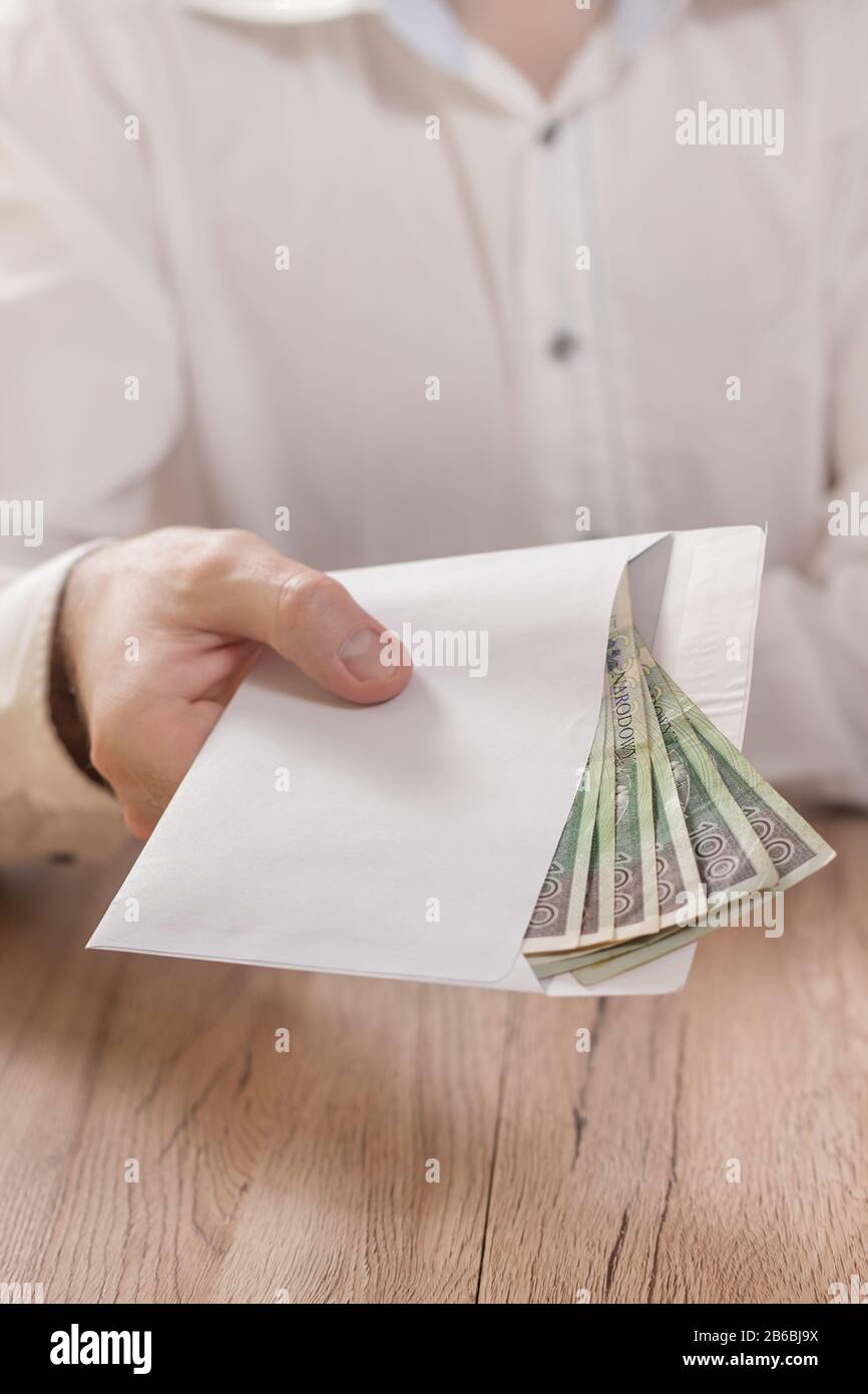 La mano di un uomo tiene una busta bianca riempita di banconote della valuta polacca e la consegna sopra il tavolo. Foto Stock