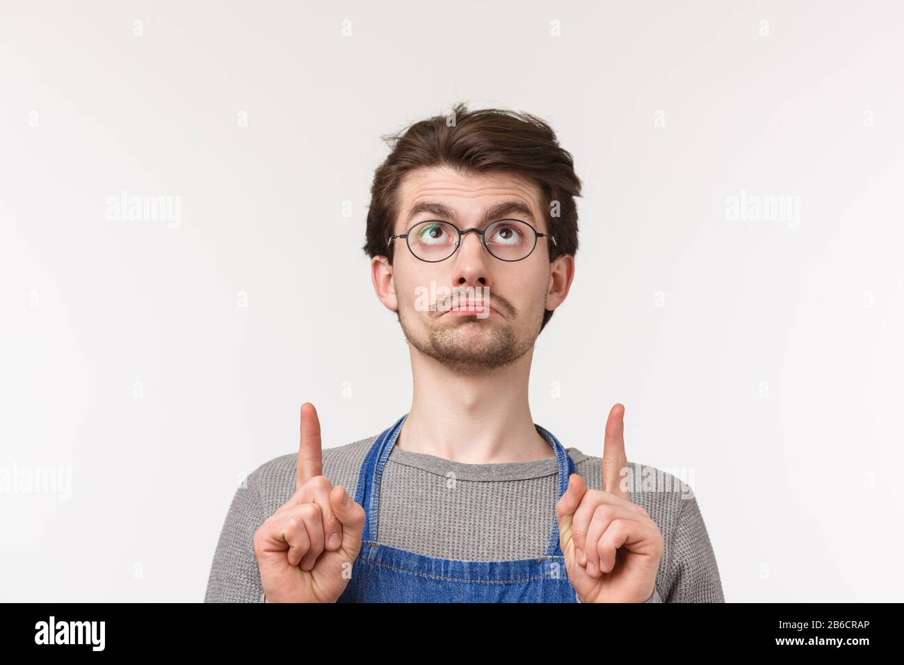 Primo piano ritratto di cupo bello bearded giovane studente maschio iniziare carriera come barista, indossare paron e occhiali, puntando le dita in su e pouting Foto Stock
