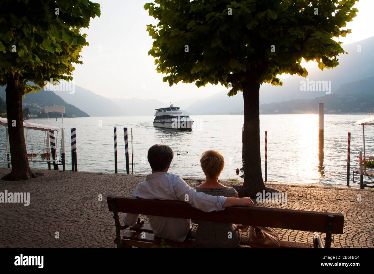 Coppia seduto su panchina e guardando traghetto che si avvicina al molo lungo il Lago di Como, Bellagio, provincia di Como, Lombardia, Italia Foto Stock