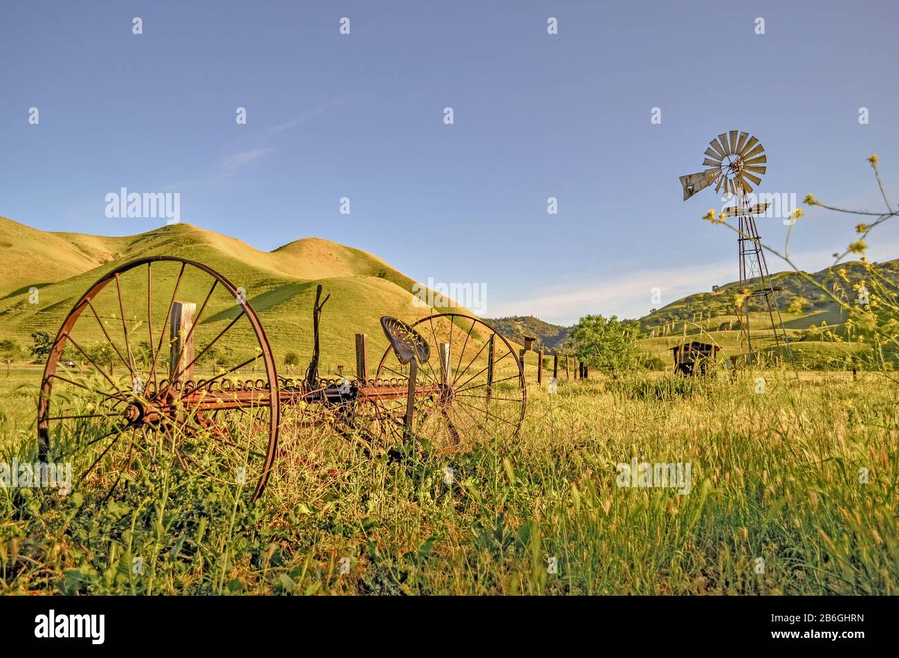 Abbandonati arrugginiti attrezzature agricole e mulino a vento con campi verdi e colline, Black Diamond Mines, Nortonville, Antioch, Pittsburg, California, Stati Uniti Foto Stock