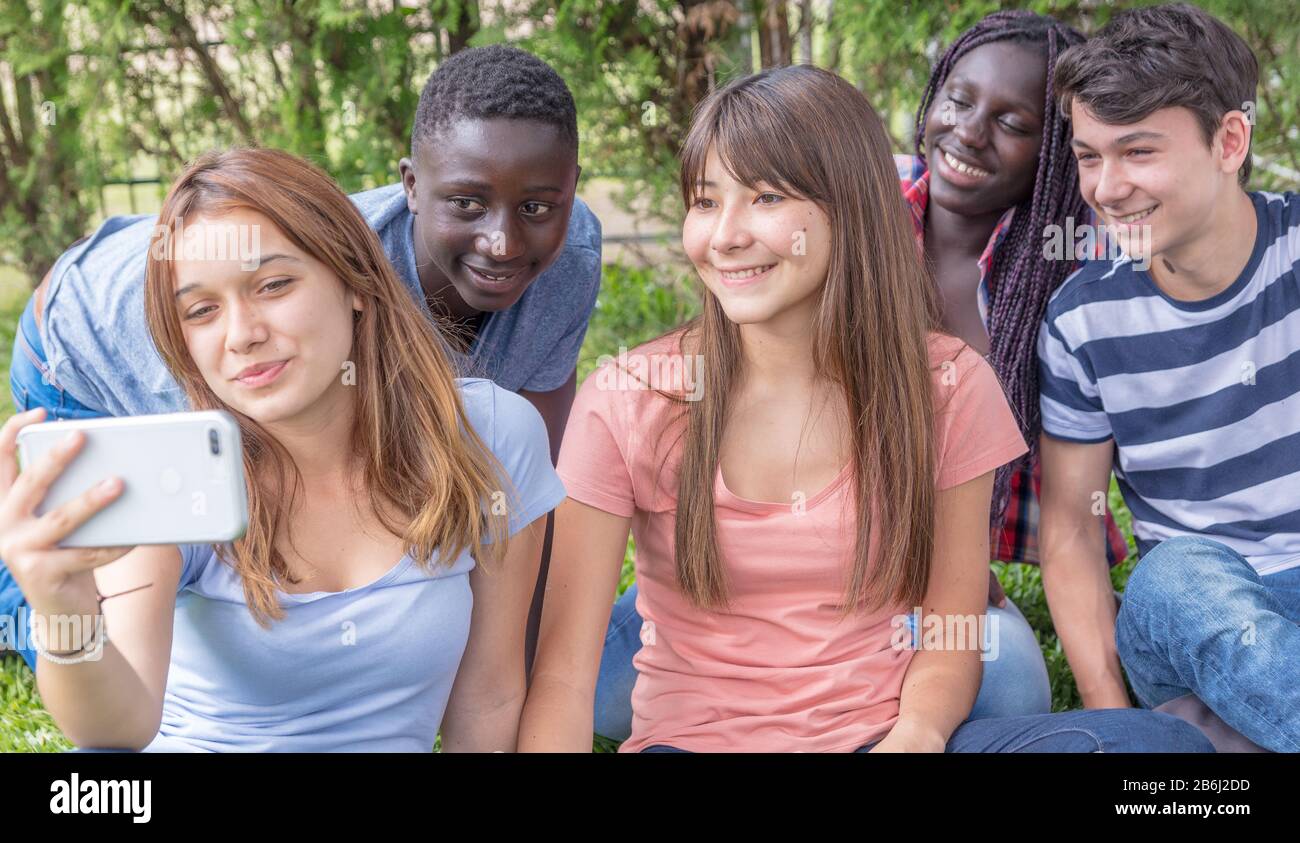 Gruppo di razze miste adolescenti palying con smarthphone sul prato facendo selfie. Foto Stock