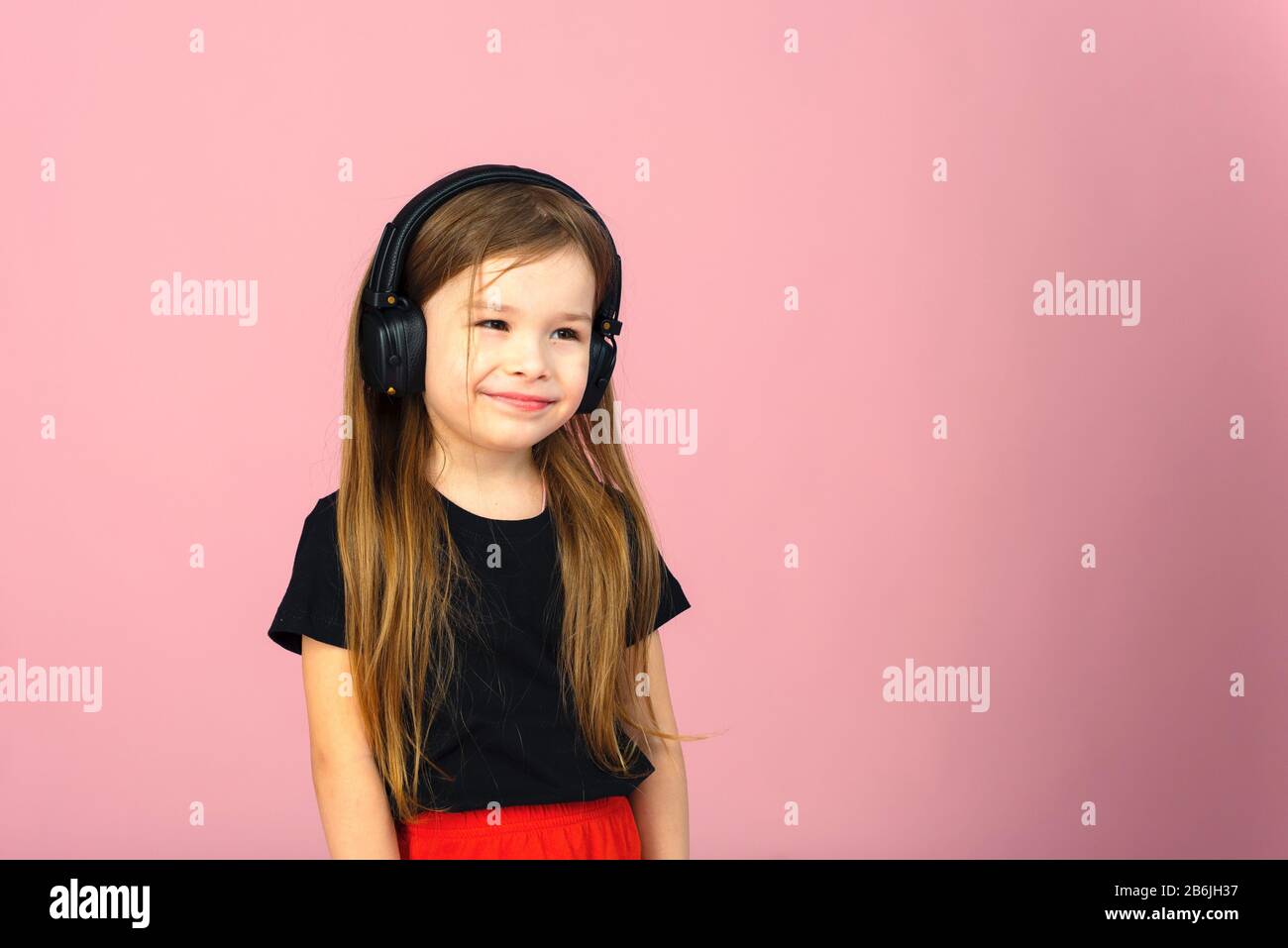 Una bambina in wireless grandi cuffie nere su uno sfondo rosa pastello guarda la fotocamera. Il concetto di ascolto e di ascolto della musica. Luogo per il testo, copyspace. Foto Stock