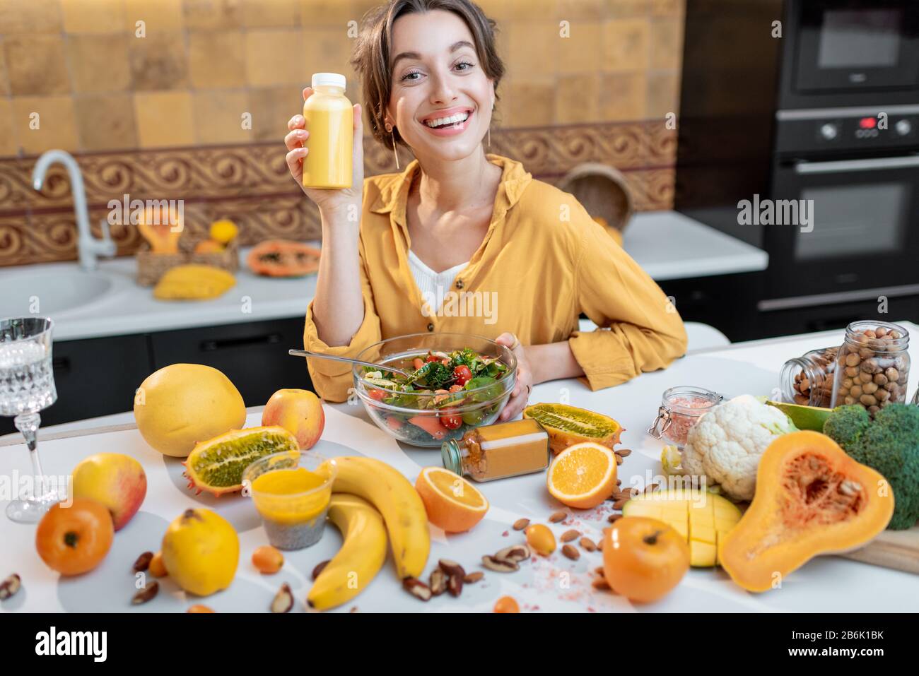 Ritratto di una giovane e gioiosa donna che tiene una bottiglia di succo al tavolo pieno di sane verdure crude e frutta. Concetto di vegetarianismo, detox, sano mangiare e benessere Foto Stock