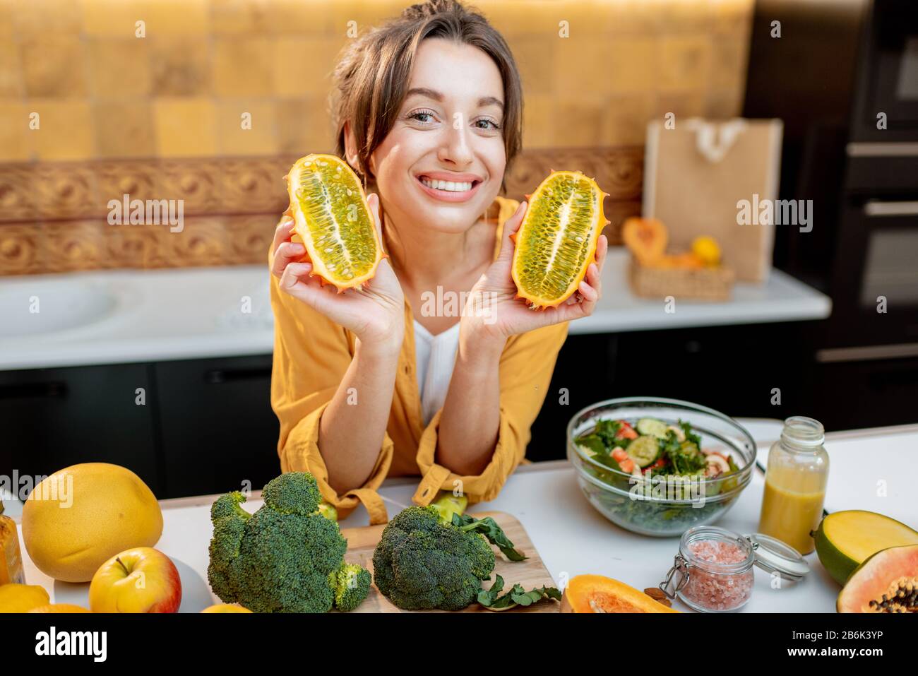 Ritratto di una donna gioiosa con varietà di frutta esotica e verdure in cucina, tenendo a fette il cetriis metuliferus. Concetto di vegetarianismo e di alimentazione sana Foto Stock