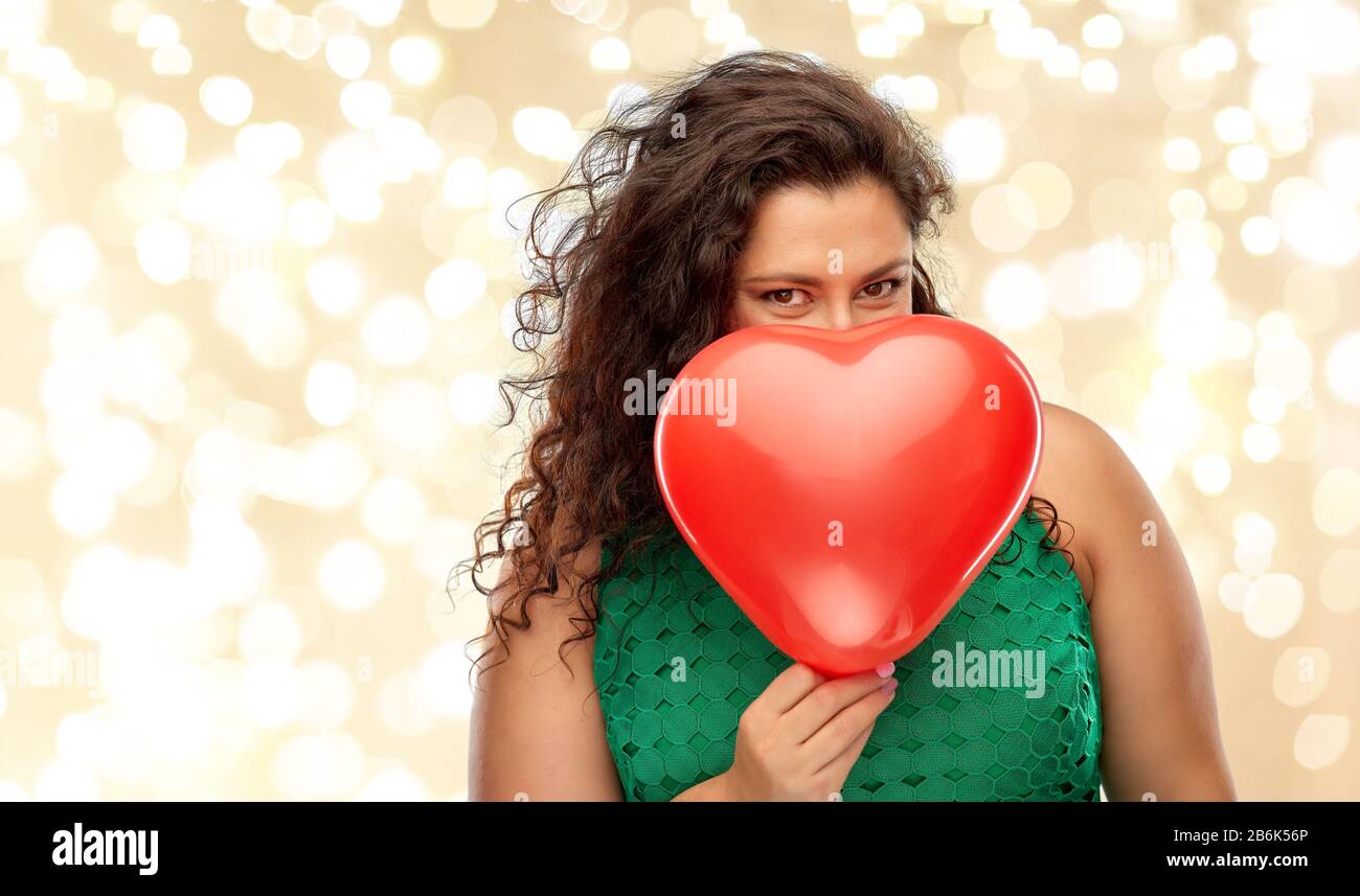 san valentino, vacanze e amore concetto - donna giocosa in abito verde nascosto dietro il cuore rosso palloncino a forma di cuore sulle luci festive su beige backgrou Foto Stock