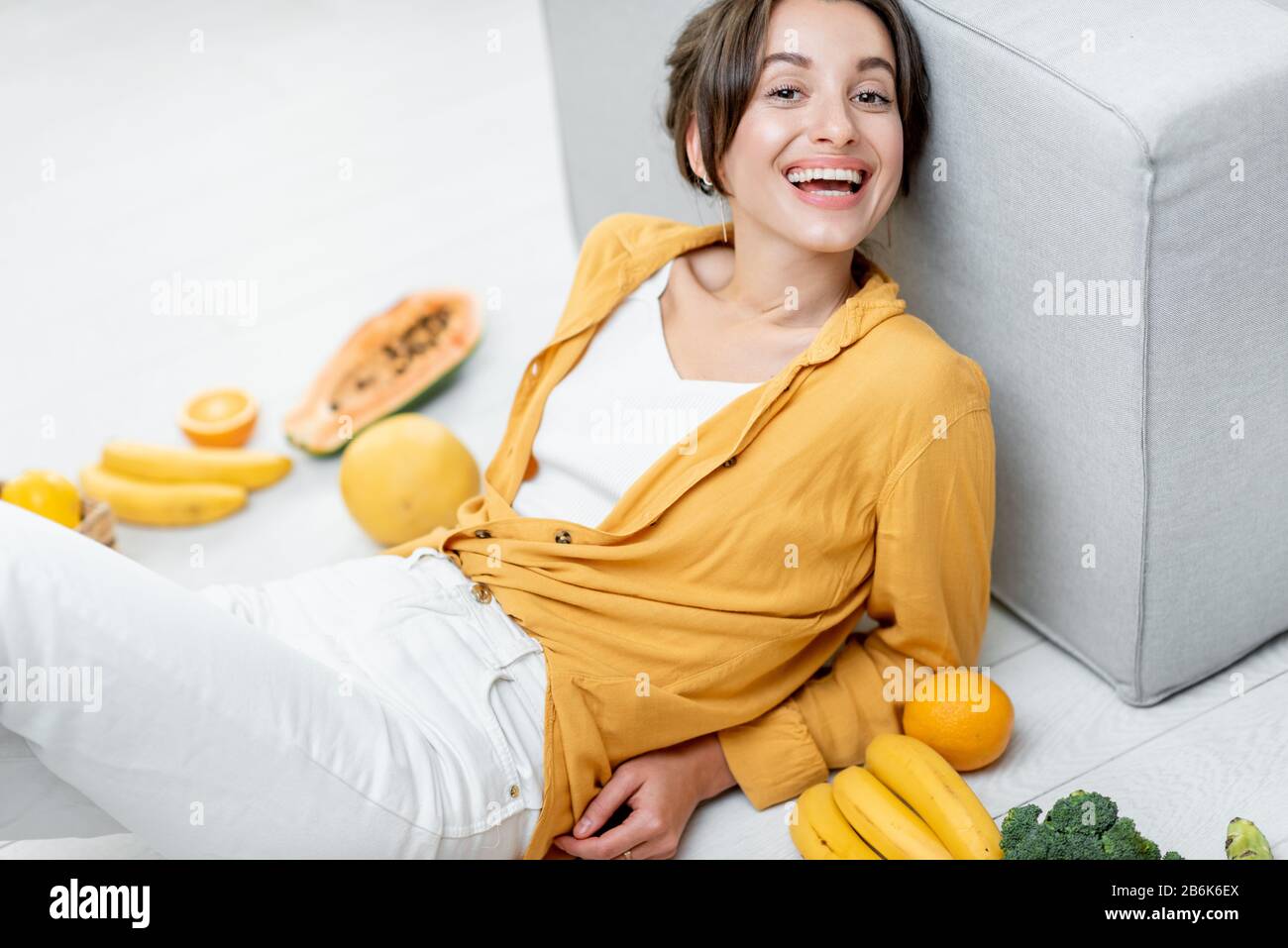 Ritratto di una giovane e allegra donna seduta con un sacco di frutta fresca e verdure sul pavimento a casa. Foto in giallo. Concetto di benessere, cibo sano e solitudine Foto Stock