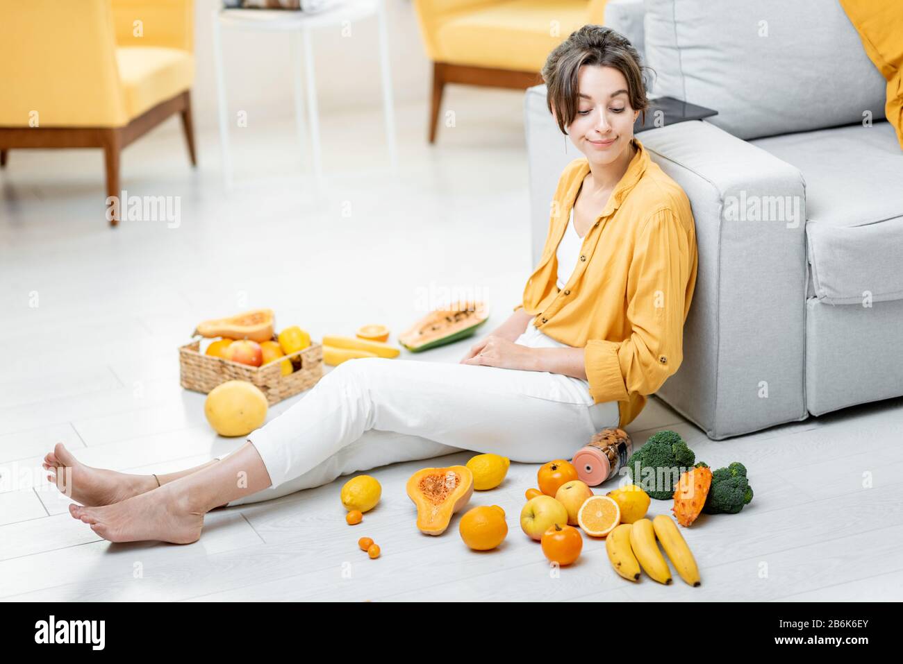 Ritratto di una giovane e allegra donna seduta con un sacco di frutta fresca e verdure sul pavimento a casa. Foto in giallo. Concetto di benessere, cibo sano e solitudine Foto Stock