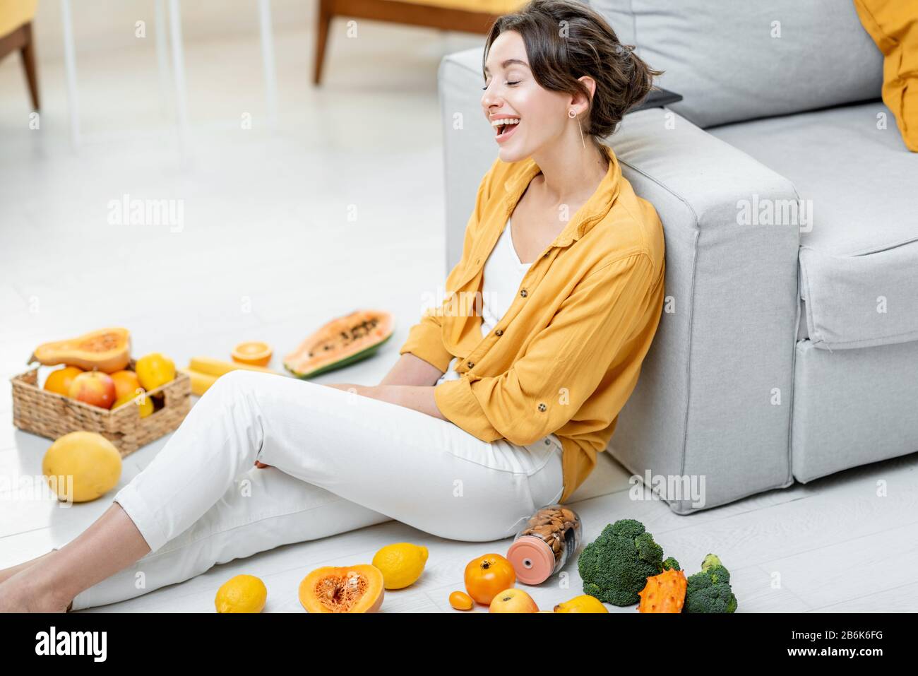 Ritratto di una giovane e allegra donna seduta con un sacco di frutta fresca e verdure sul pavimento a casa. Foto in giallo. Concetto di benessere, cibo sano e solitudine Foto Stock