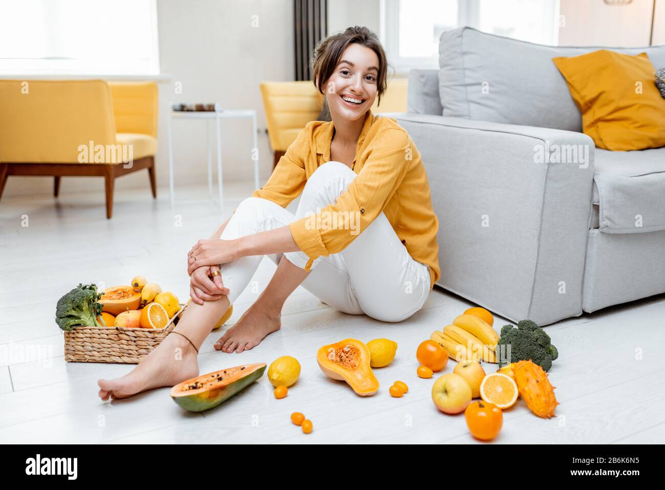 Ritratto di una giovane e allegra donna seduta con un sacco di frutta fresca e verdure sul pavimento a casa. Foto in giallo. Concetto di benessere, cibo sano e solitudine Foto Stock