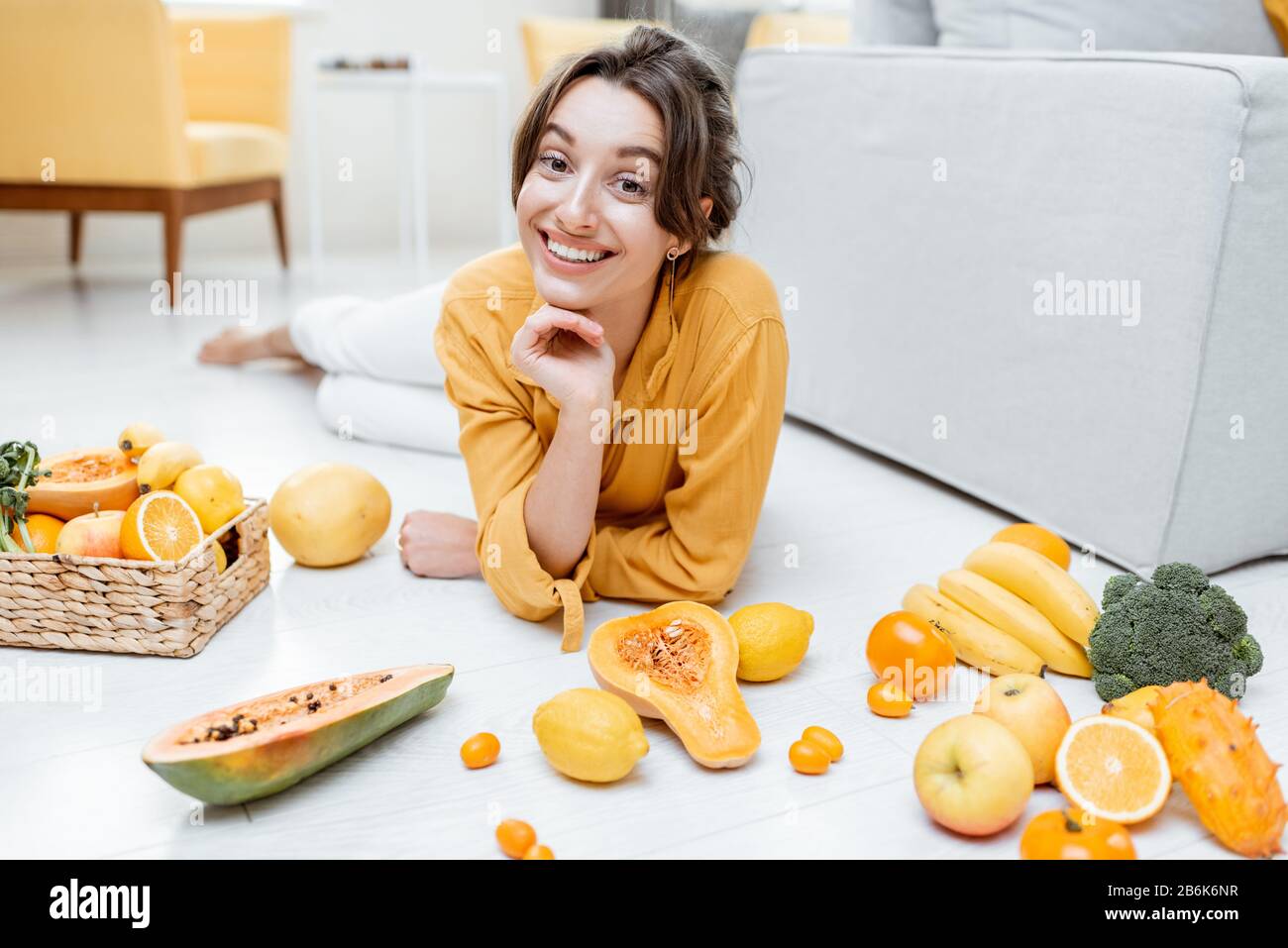 Ritratto di una giovane e allegra donna sdraiata con un sacco di frutta fresca e verdure sul pavimento a casa. Foto in giallo. Concetto di benessere, cibo sano e solitudine Foto Stock
