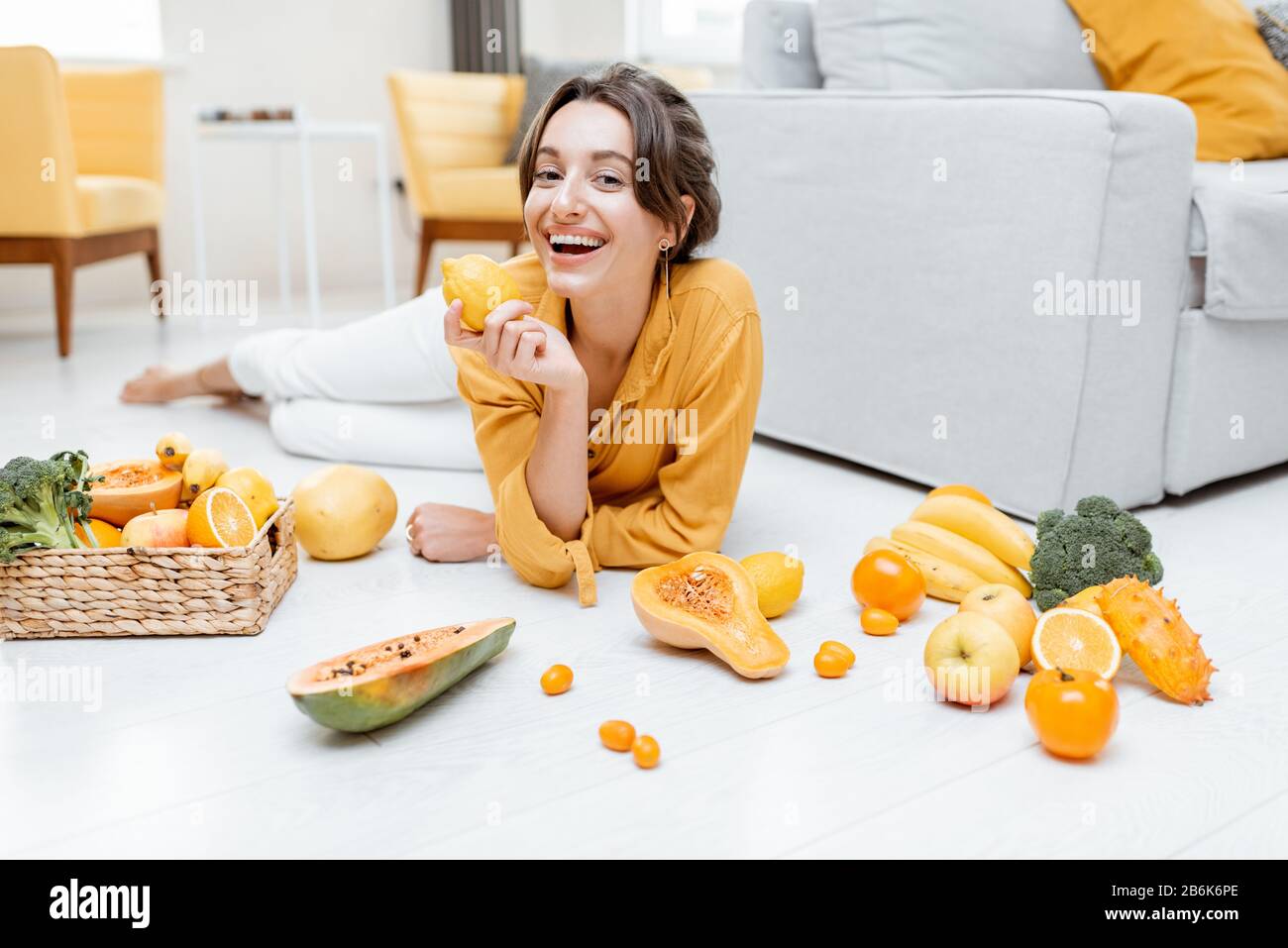 Ritratto di una giovane e allegra donna sdraiata con un sacco di frutta fresca e verdure sul pavimento a casa. Foto in giallo. Concetto di benessere, cibo sano e solitudine Foto Stock