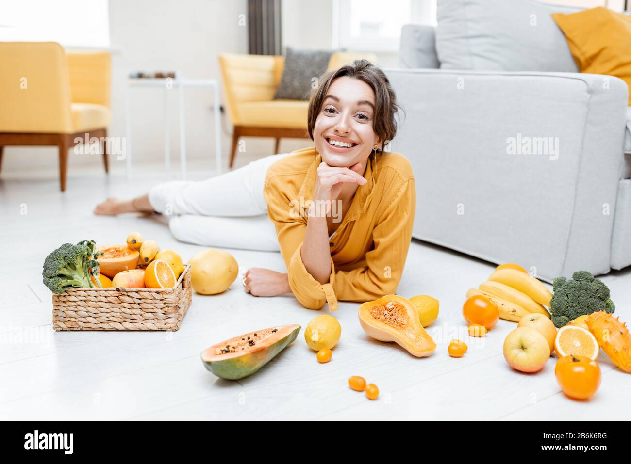 Ritratto di una giovane e allegra donna sdraiata con un sacco di frutta fresca e verdure sul pavimento a casa. Foto in giallo. Concetto di benessere, cibo sano e solitudine Foto Stock