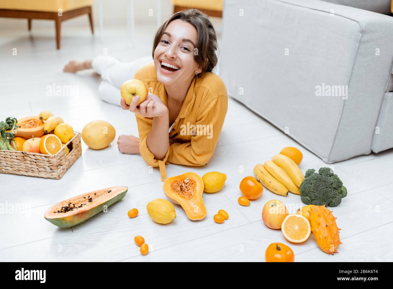 Ritratto di una giovane e allegra donna sdraiata con un sacco di frutta fresca e verdure sul pavimento a casa. Foto in giallo. Concetto di benessere, cibo sano e solitudine Foto Stock