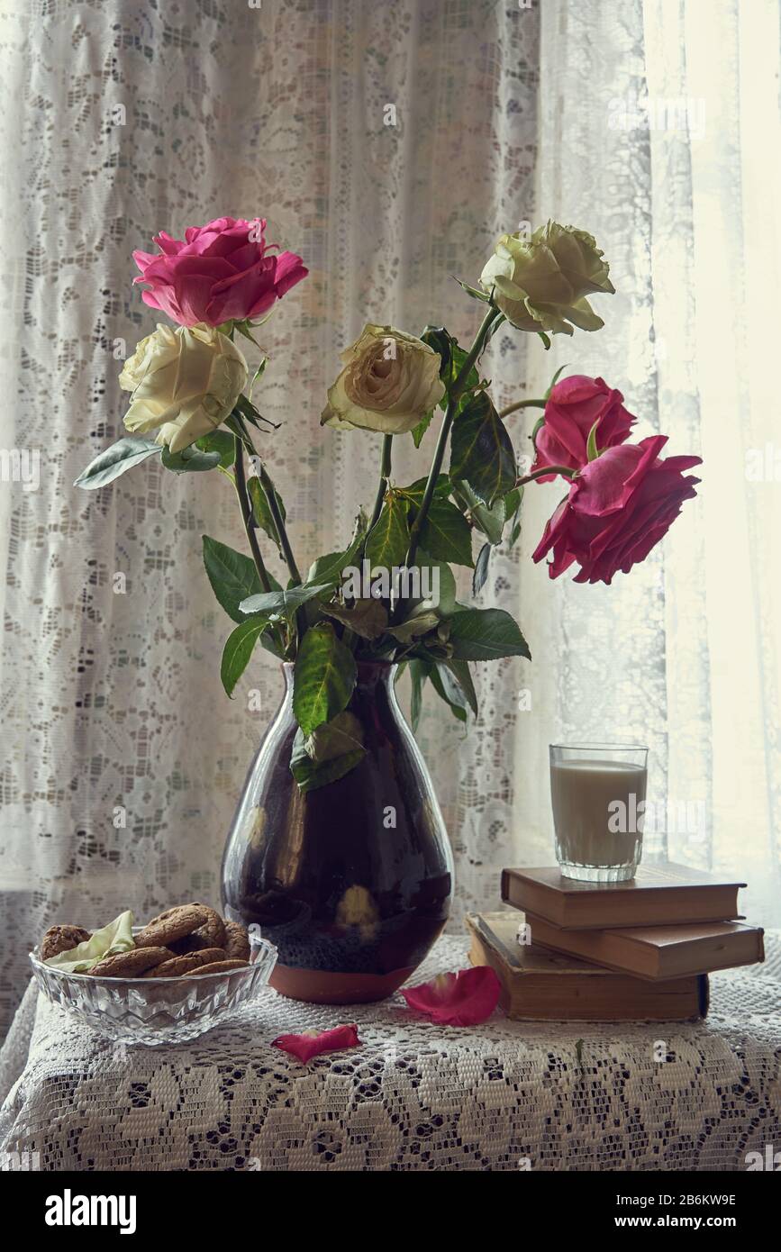 Un bouquet di rose in vaso di ceramica . Accanto ad esso ci sono libri , un bicchiere di latte, e una ciotola di biscotti. Foto Stock