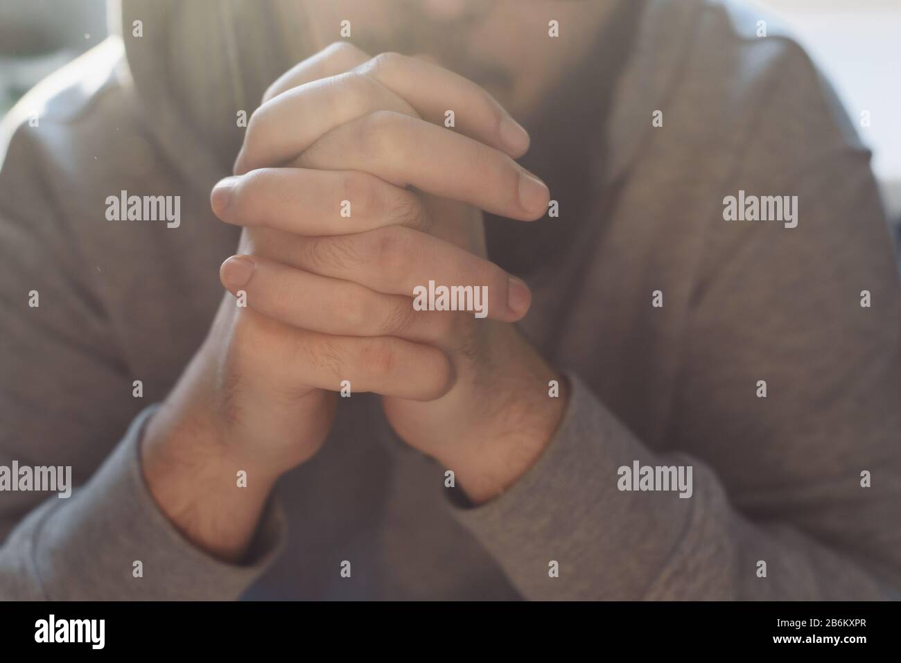 Uomo adulto bearded che prega a Dio che siede a casa nel raggio del sole. Un musulmano o cristiano alza le mani a Dio. Mani incrociate nel gesto di preghiera da vicino. Foto Stock