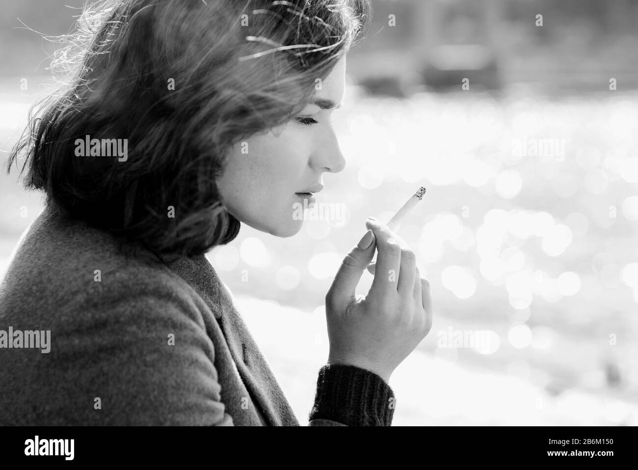 bella donna giovane, trista e stressante, in maglione e cappotto fumando sigaretta vicino al fiume Foto Stock