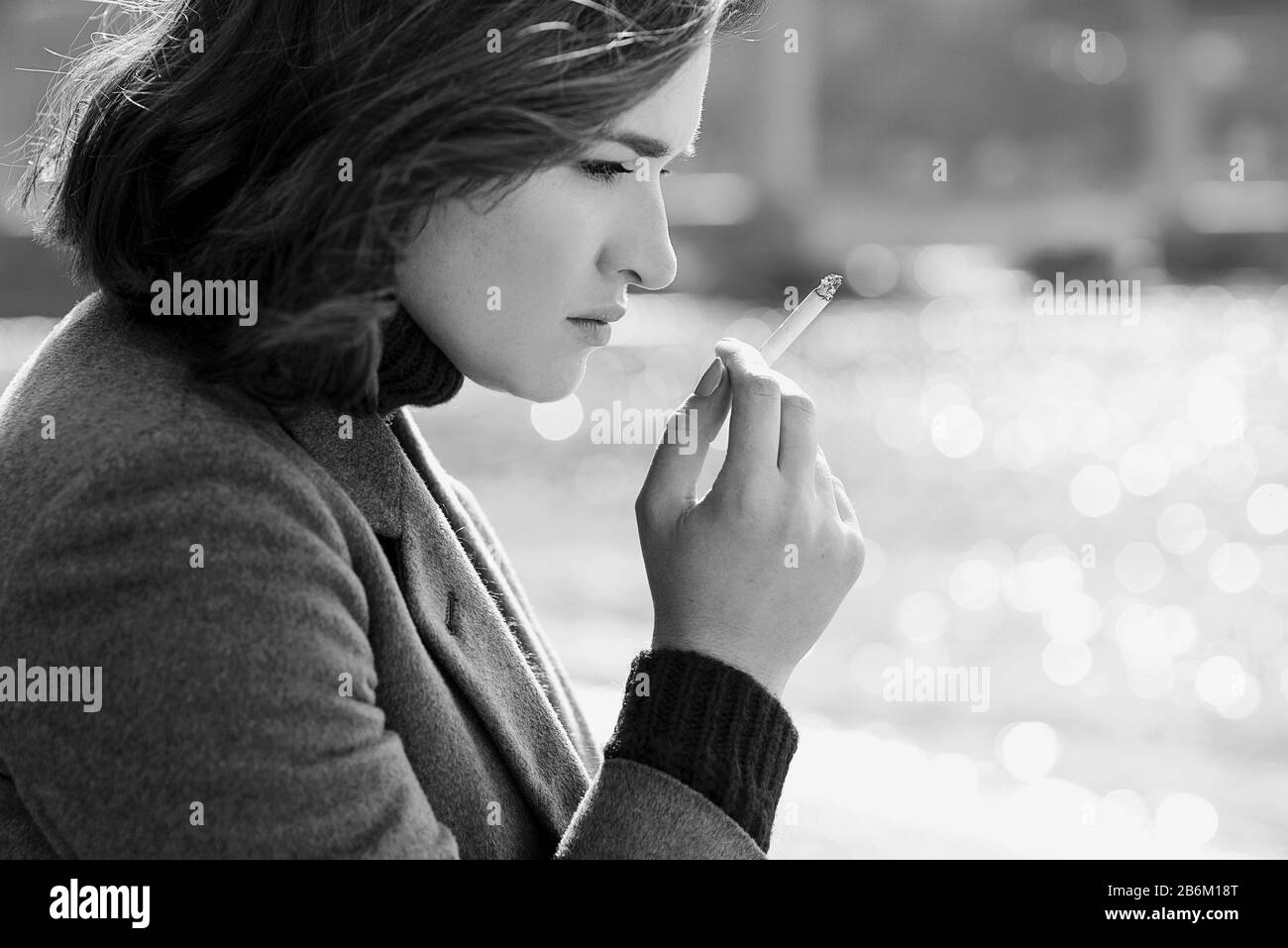 bella donna giovane, trista e stressante, in maglione e cappotto fumando sigaretta vicino al fiume Foto Stock