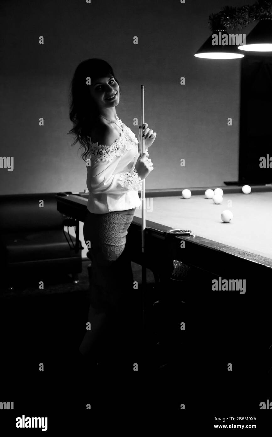 giovane donna con capelli neri in una blusa bianca si alza vicino ad un poo,l tavolo tiene un cue nelle sue mani, Foto Stock