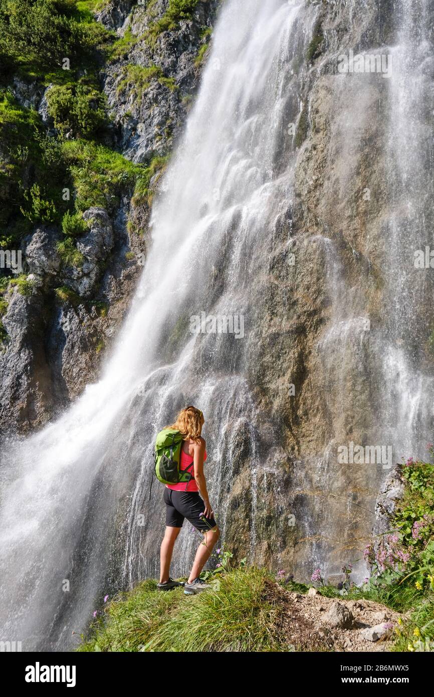 Donna escursionista, con uno zaino verde, guarda la cascata Dalfazer, Austria. Foto Stock